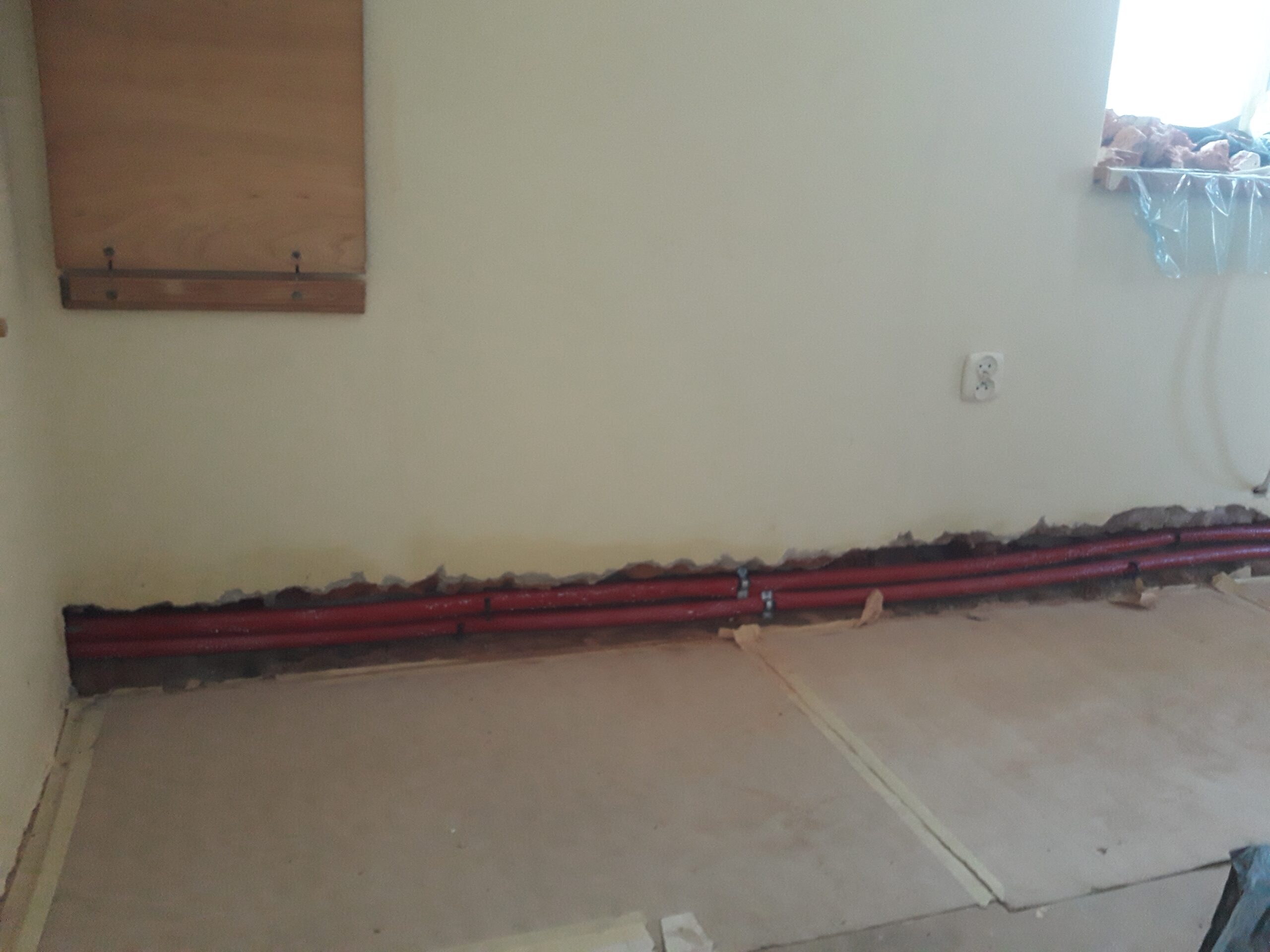 Odcinek instalacji grzewczej poprowadzonej wzdłuż ściany w pomieszczeniu podczas remontu, z zabezpieczoną podłogą i przygotowaniem do dalszych prac.