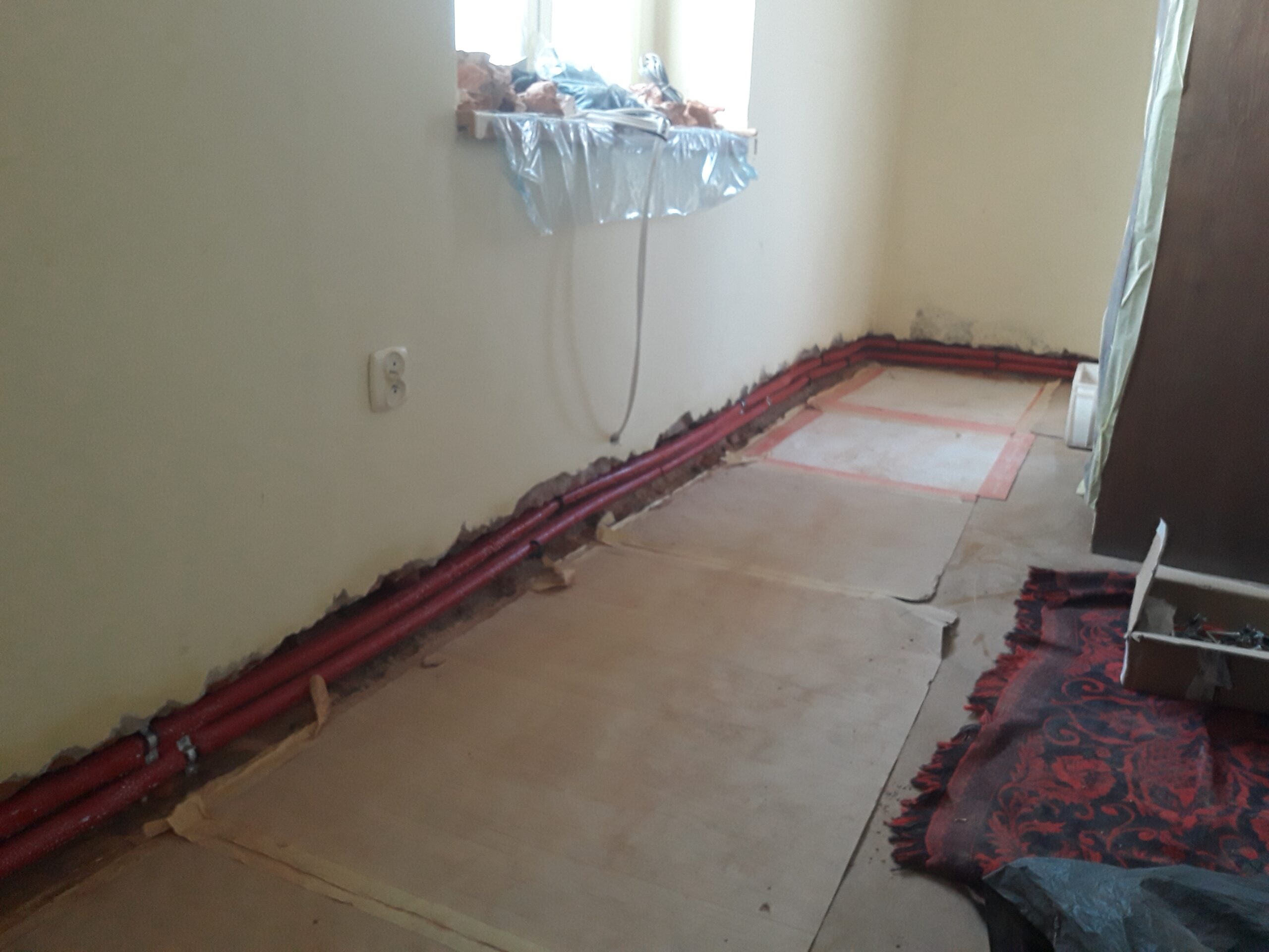 Pomieszczenie w trakcie remontu z poprowadzoną instalacją grzewczą wzdłuż ściany, zabezpieczoną podłogą i przygotowaniem do kolejnych prac wykończeniowych.