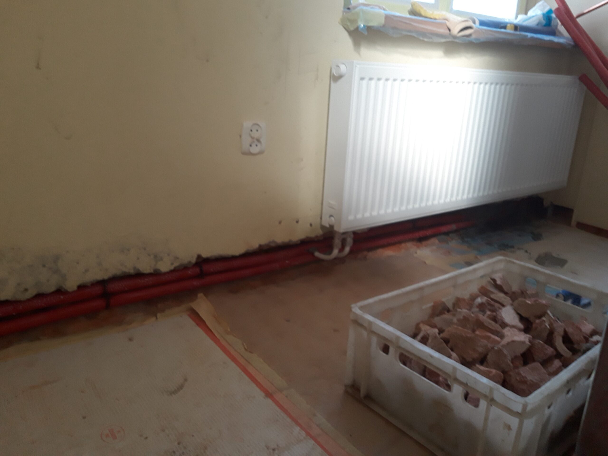 Grzejnik ścienny z poprowadzoną instalacją grzewczą przy podłodze w pomieszczeniu podczas prac remontowych, z widocznymi materiałami przygotowanymi do dalszych etapów.