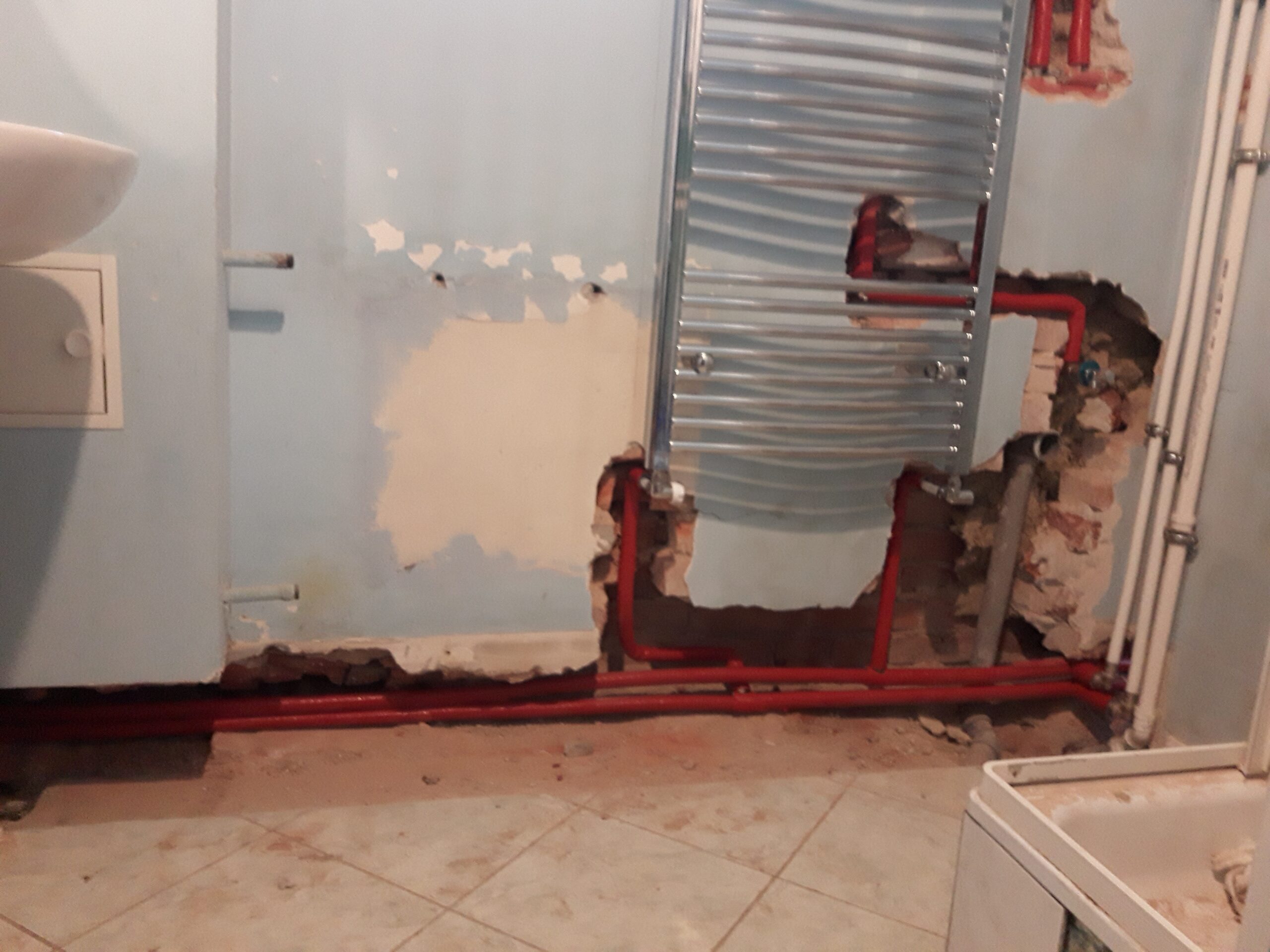 Ściana łazienki w trakcie remontu z odsłoniętą instalacją grzewczą, zamontowanym grzejnikiem drabinkowym i przygotowaniem do dalszych prac wykończeniowych.