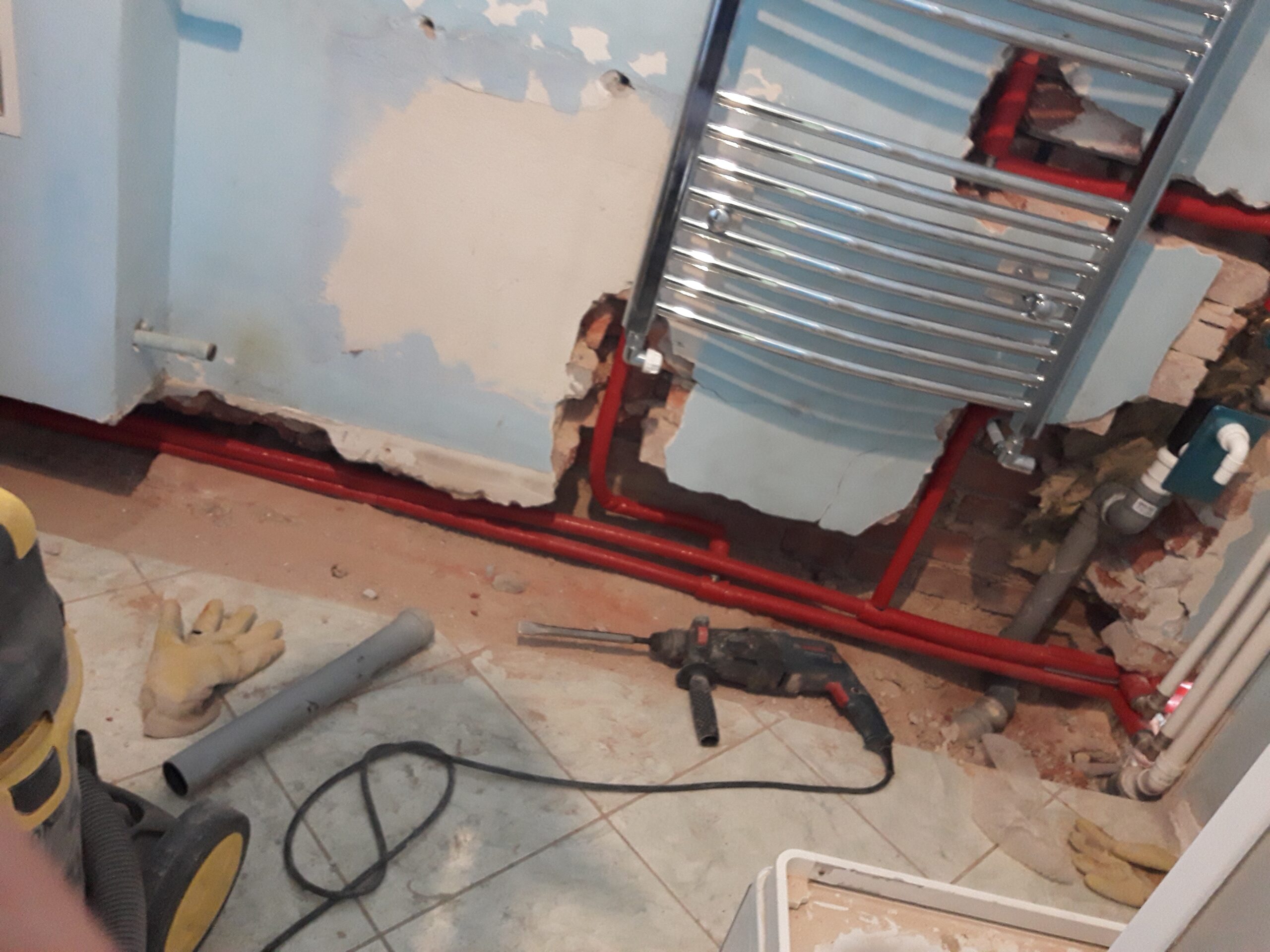 Fragment łazienki w trakcie prac instalacyjnych z odsłoniętymi rurami grzewczymi, grzejnikiem drabinkowym oraz narzędziami używanymi podczas remontu.