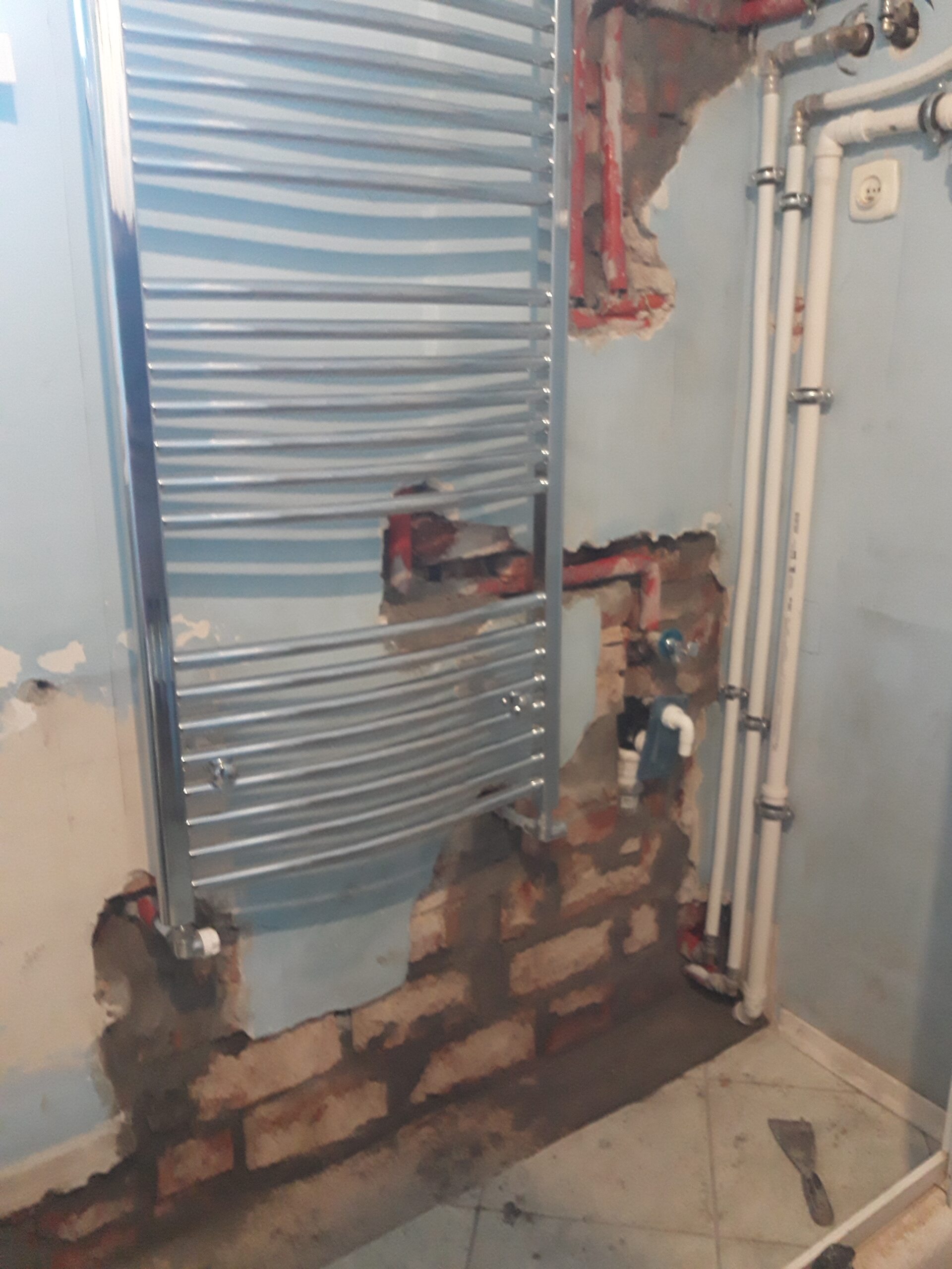 Zbliżenie na grzejnik drabinkowy oraz odsłoniętą instalację wodną i grzewczą w łazience, pokazujące etap prac remontowych przed wykończeniem ścian.