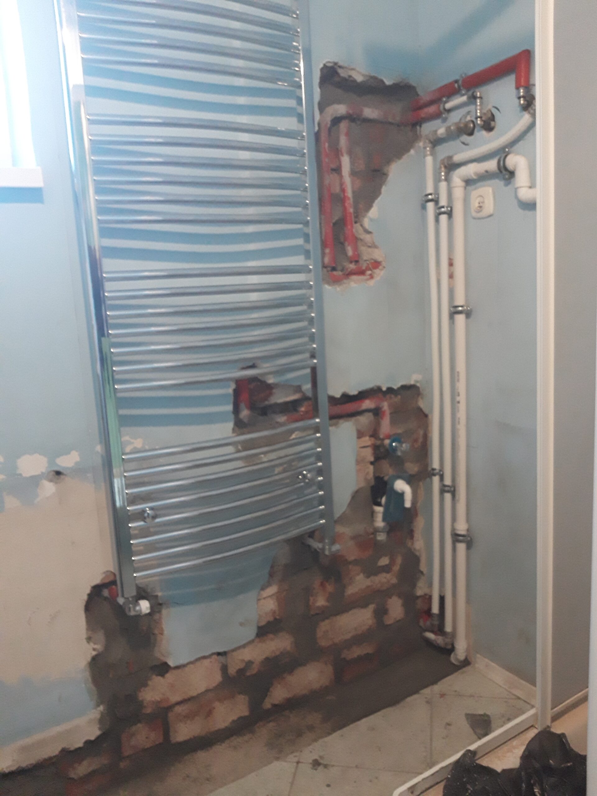 Fragment łazienki w trakcie remontu z widoczną instalacją wodną i grzewczą oraz grzejnikiem drabinkowym zamontowanym na nieukończonej ścianie.