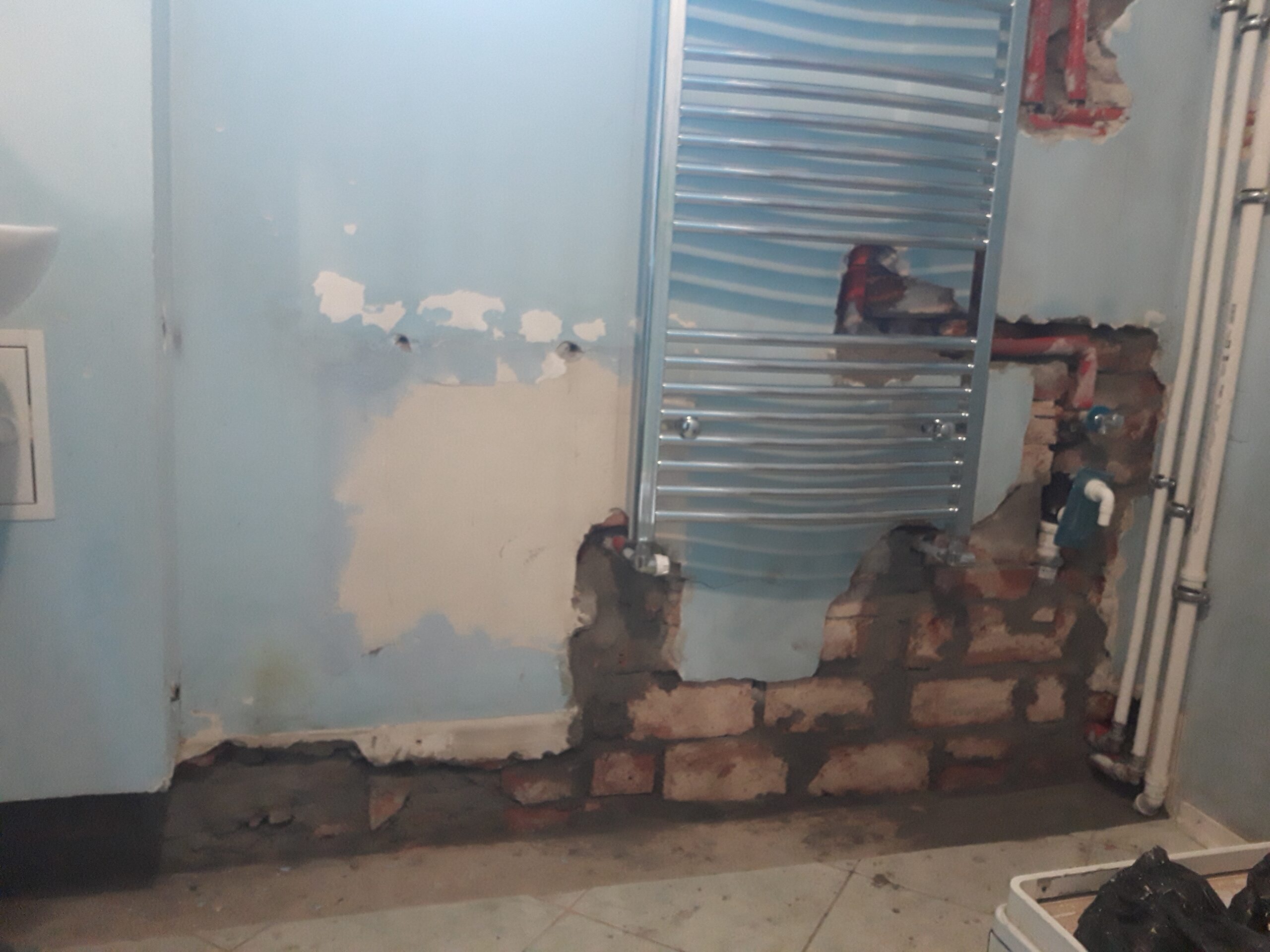 Ściana łazienki w trakcie remontu z odsłoniętą instalacją, grzejnikiem drabinkowym i fragmentami muru przygotowanymi do dalszych prac wykończeniowych.
