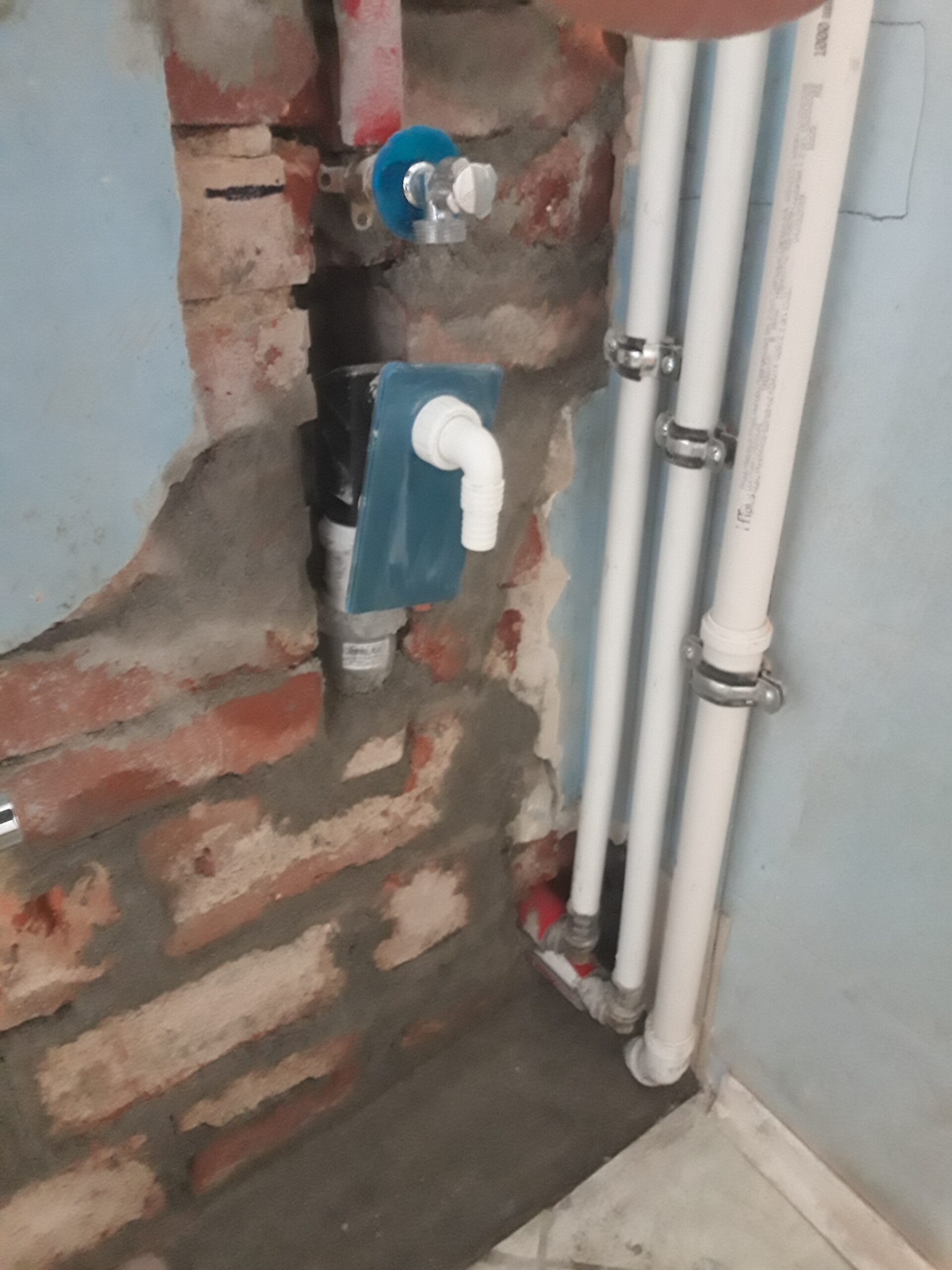 Odsłonięta instalacja wodno-kanalizacyjna w ścianie z widocznymi rurami, zaworem i elementami podłączenia, pokazująca etap prac remontowych.