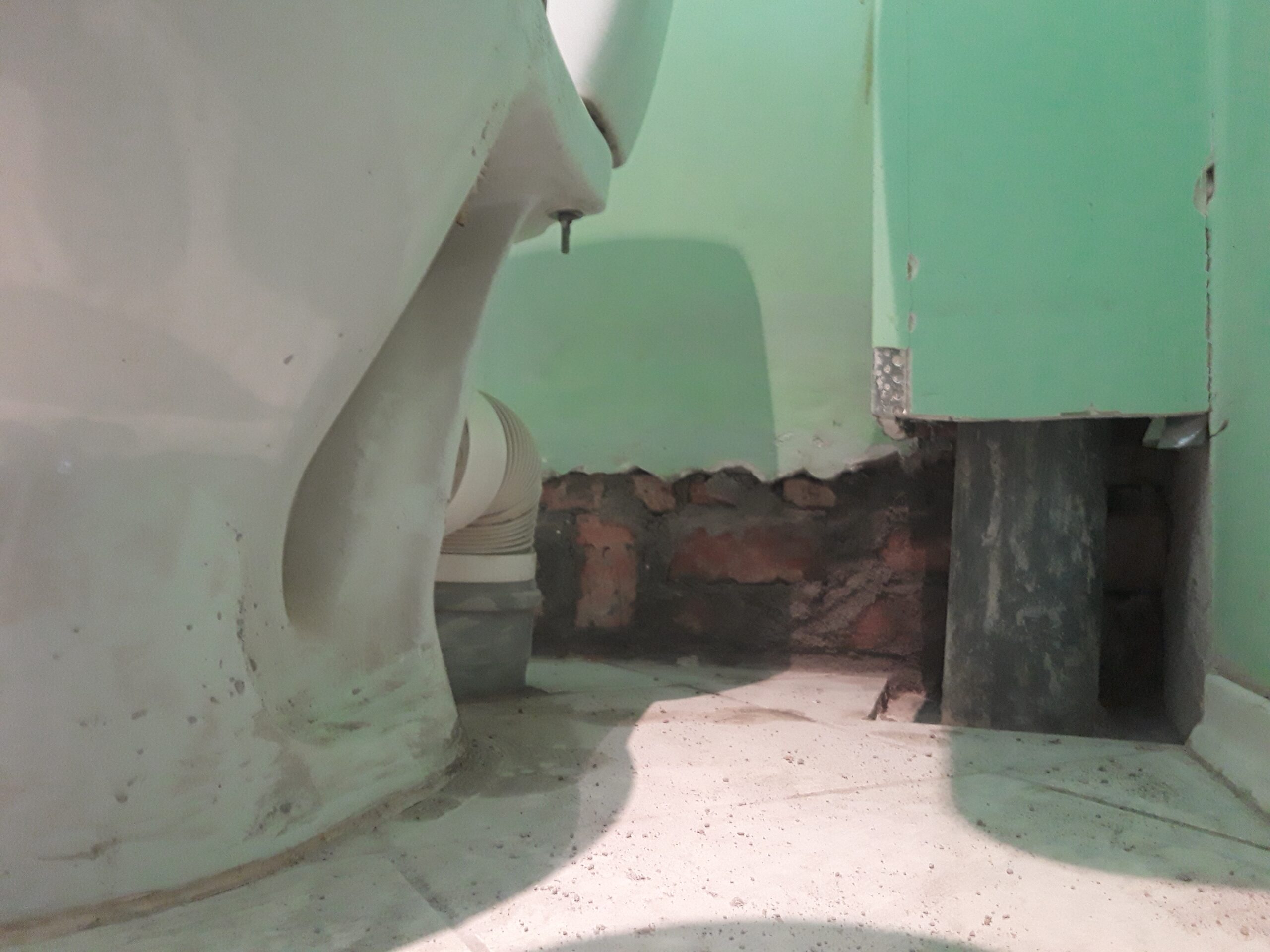 Dolna część toalety z odsłoniętą ścianą i elementami instalacji, pokazująca etap prac remontowych przy podłączeniach sanitarnych.