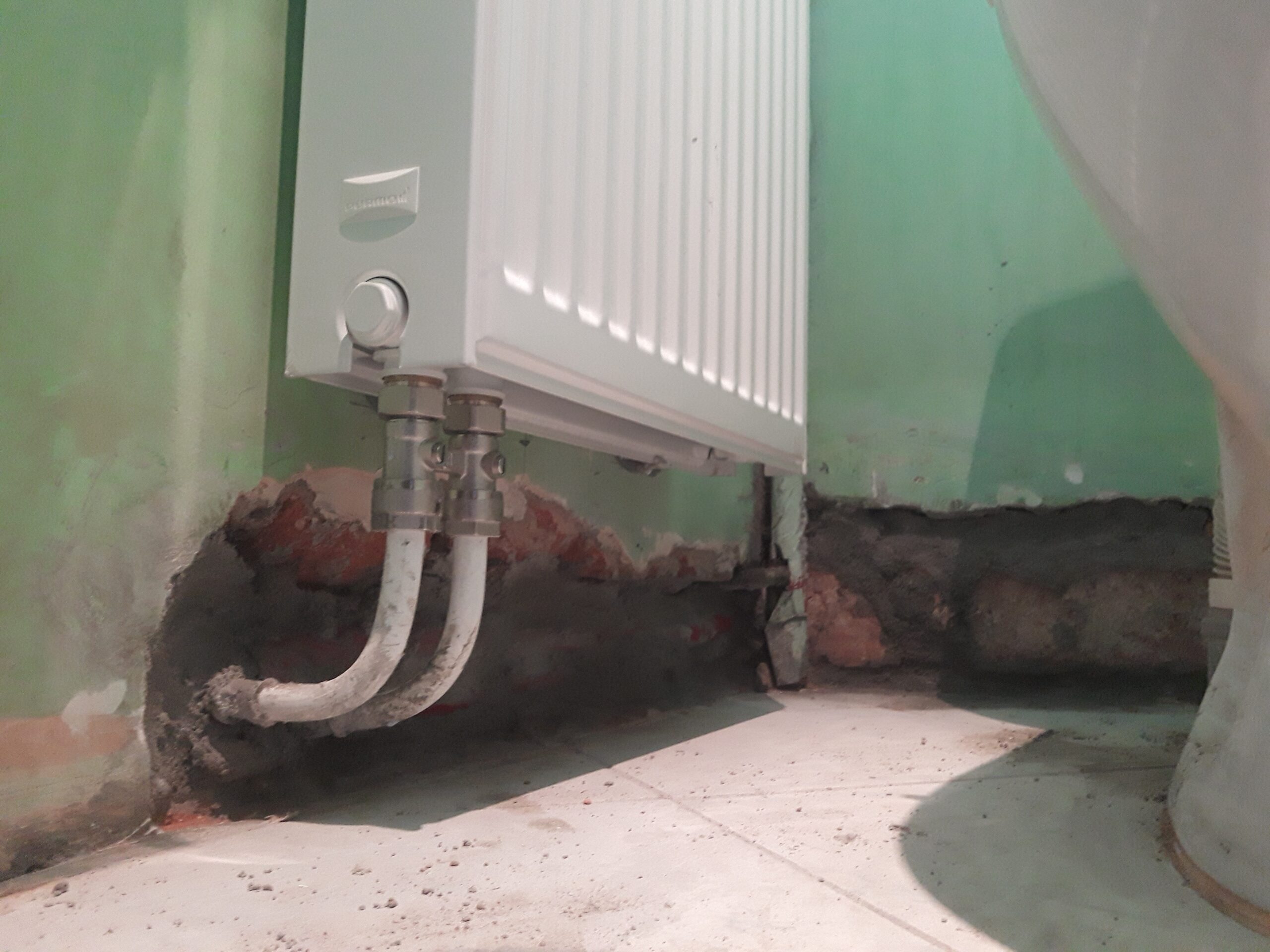 Dolna część grzejnika ściennego z podłączonymi rurami instalacyjnymi, pokazująca etap prac remontowych przy odsłoniętej ścianie i podłodze.