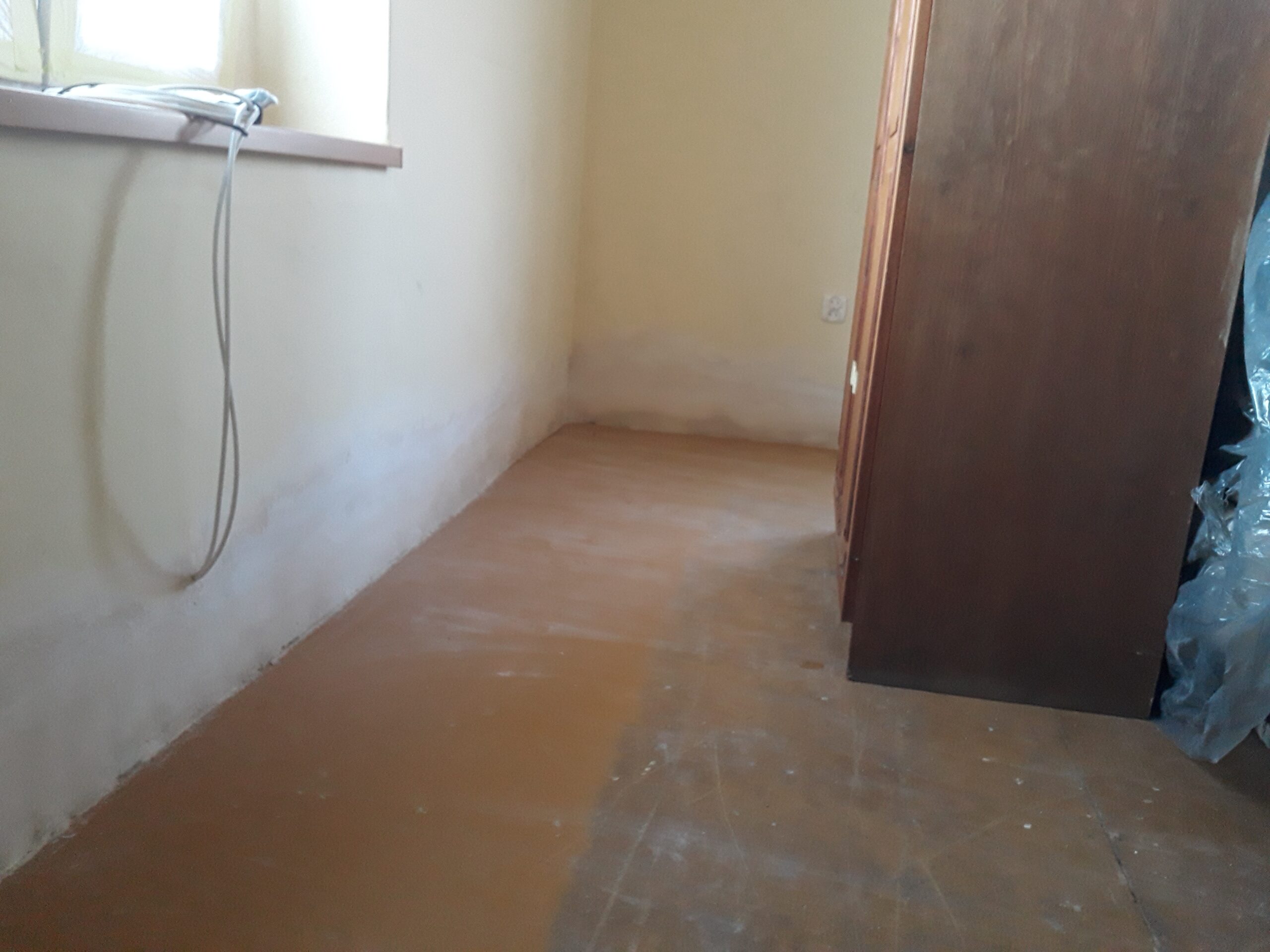 Puste pomieszczenie z wyrównaną podłogą przed dalszymi pracami wykończeniowymi, z widocznym oknem, ścianami i meblami odsuniętymi na bok.