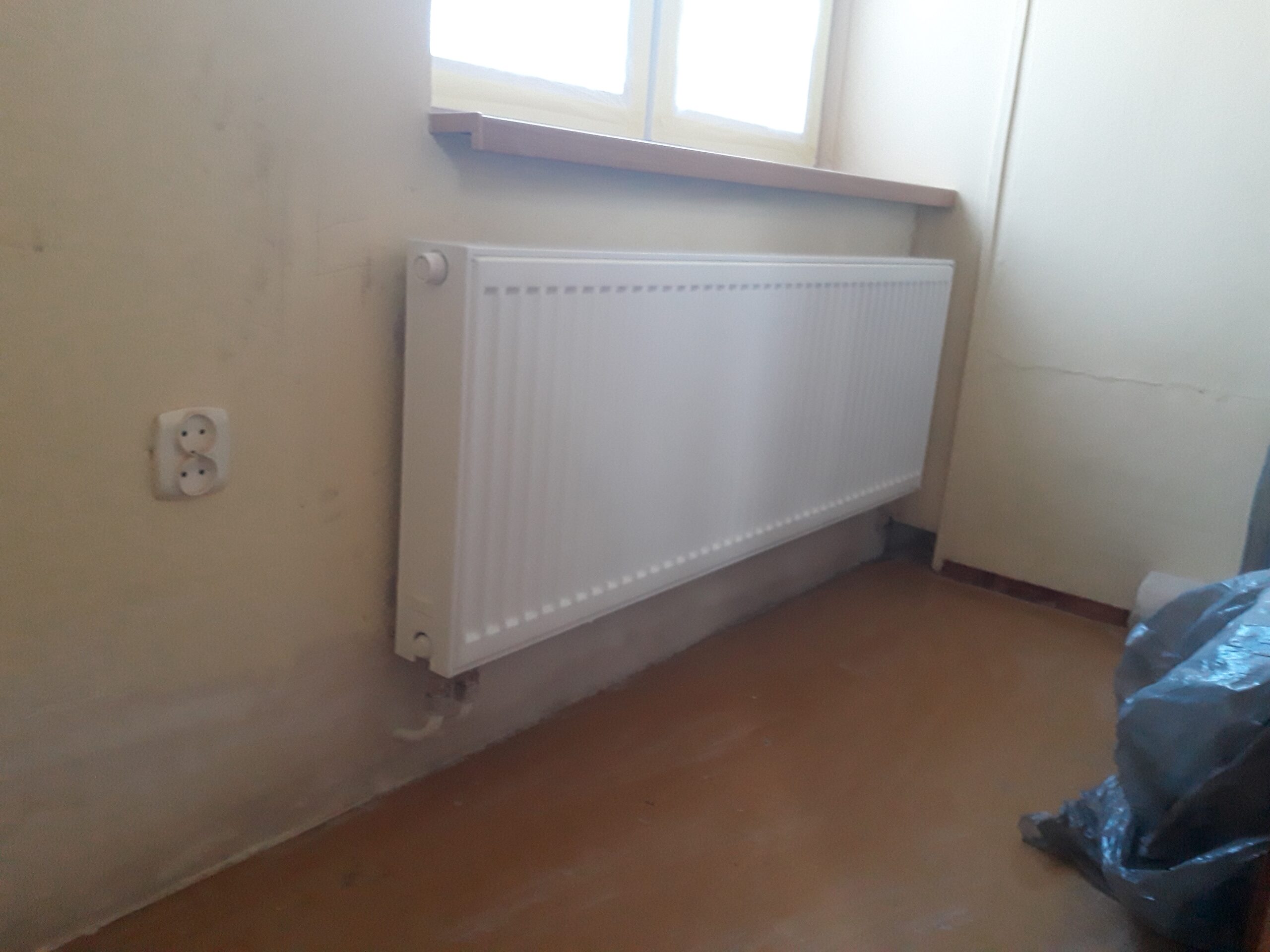 Zamontowany grzejnik ścienny pod oknem w pomieszczeniu w trakcie prac wykończeniowych, z widoczną surową podłogą i nieukończonymi ścianami.