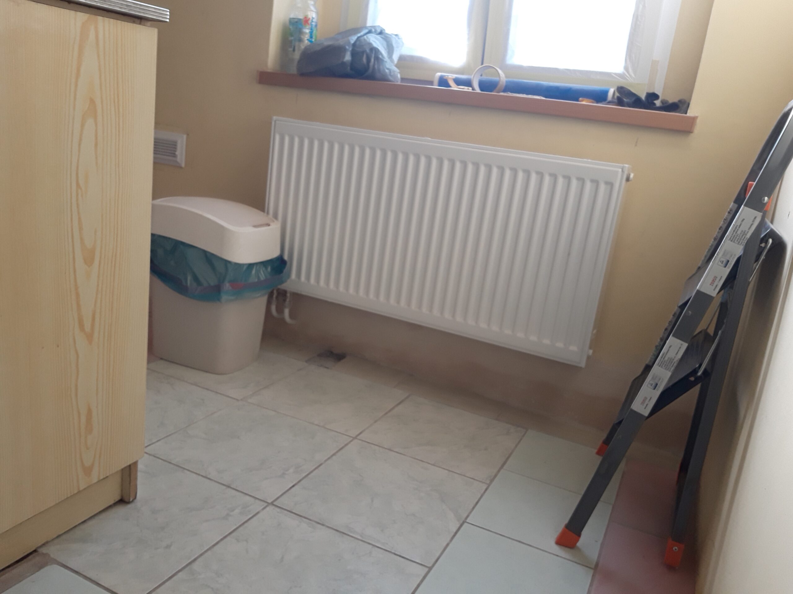 Fragment kuchni po remoncie z ułożoną podłogą z płytek, zamontowanym grzejnikiem pod oknem oraz widocznymi narzędziami używanymi podczas prac.