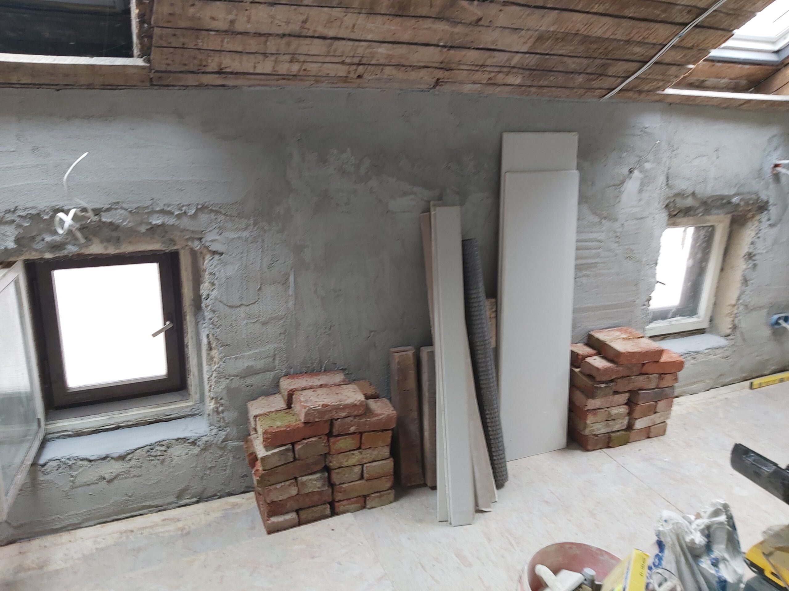 Ściana poddasza w trakcie remontu z dwoma małymi oknami, surowym tynkiem, ułożonymi cegłami oraz przygotowanymi płytami i materiałami budowlanymi do dalszych prac wykończeniowych.