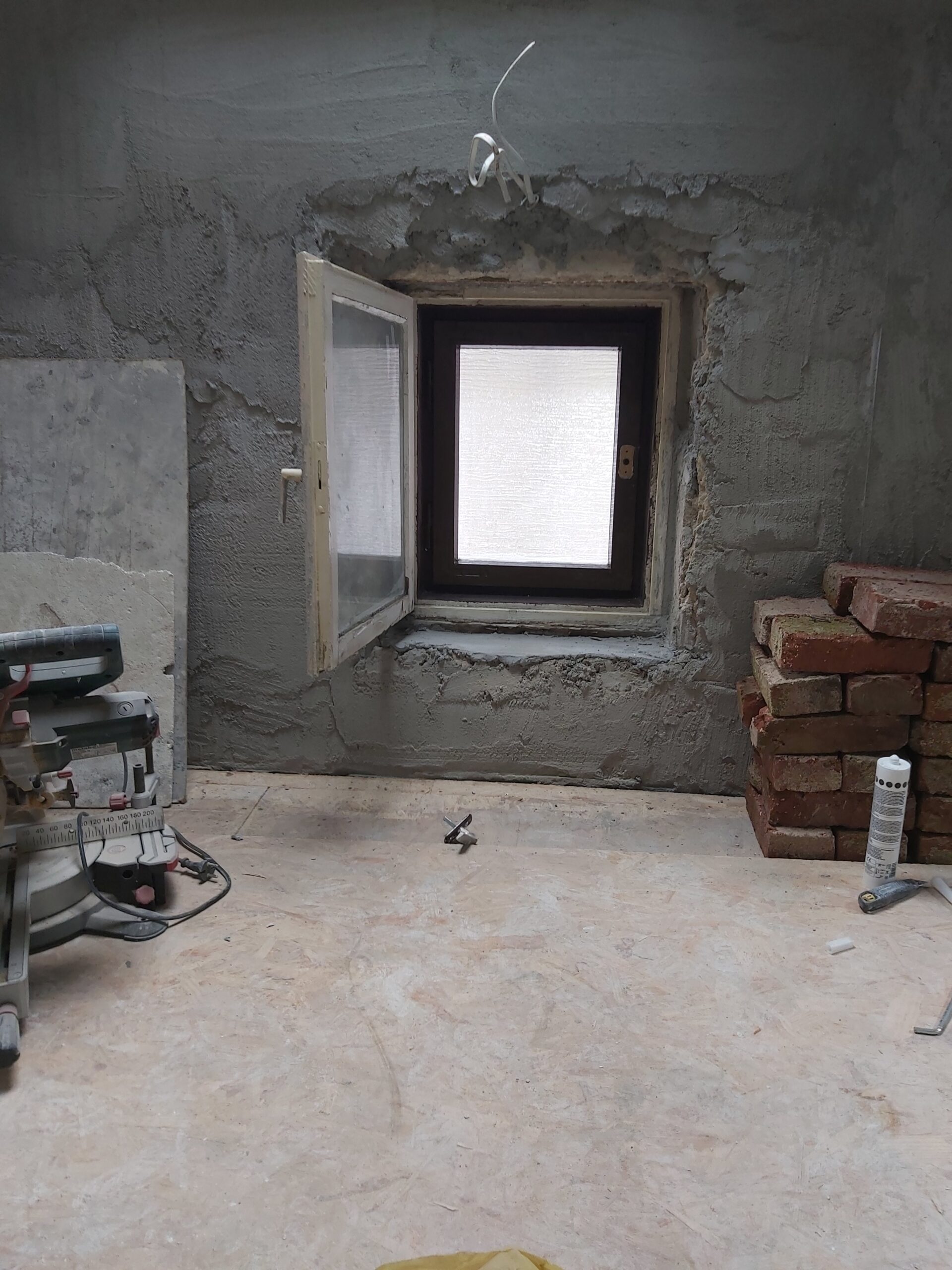 Otwarty mały lufcik w ścianie poddasza podczas remontu, surowe tynki wokół okna, cegły i narzędzia budowlane przygotowane do dalszych prac wykończeniowych.