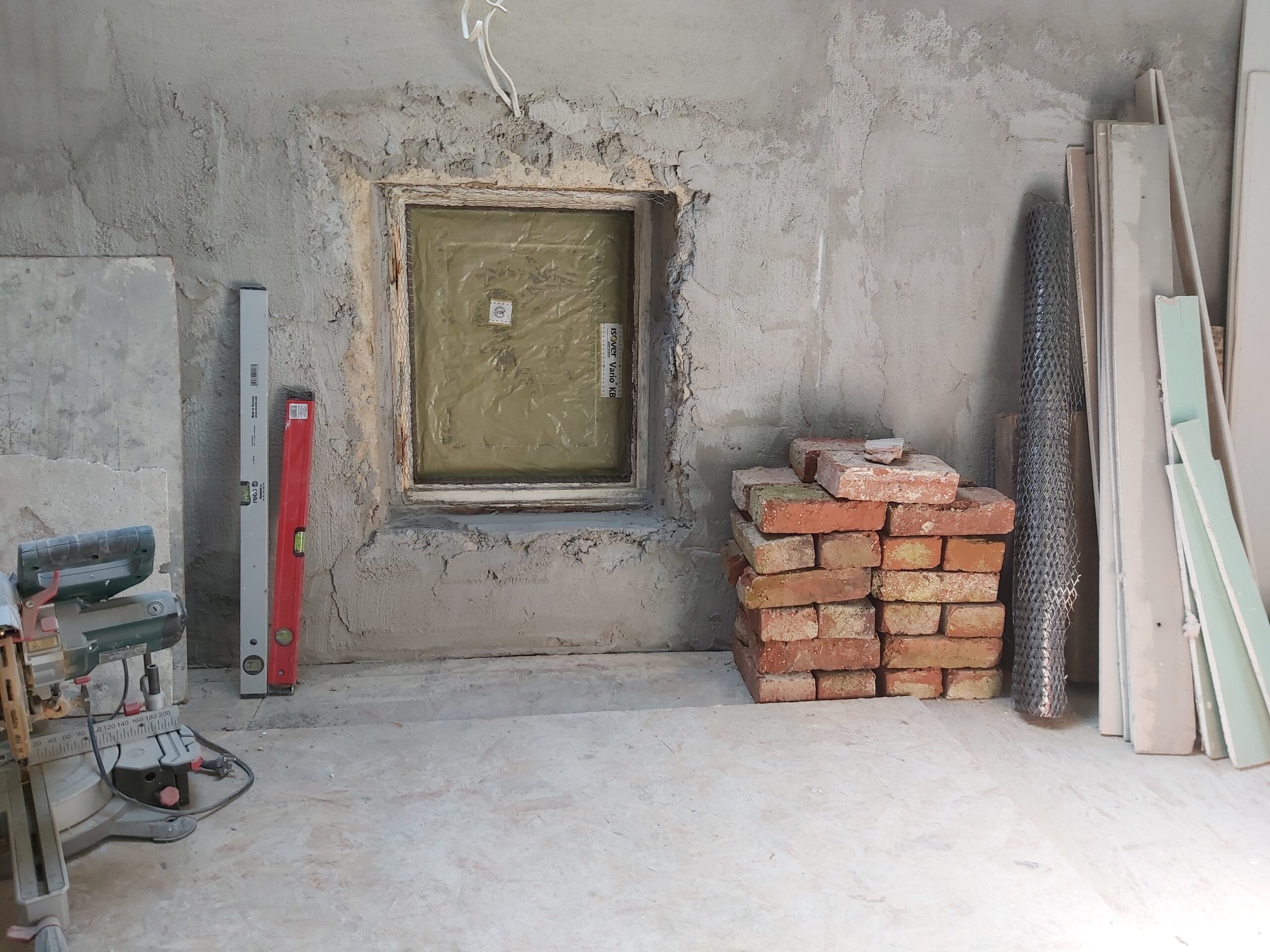 Ściana poddasza w trakcie remontu z zabezpieczonym otworem okiennym, ułożonymi cegłami oraz materiałami budowlanymi przygotowanymi do dalszych prac wykończeniowych.