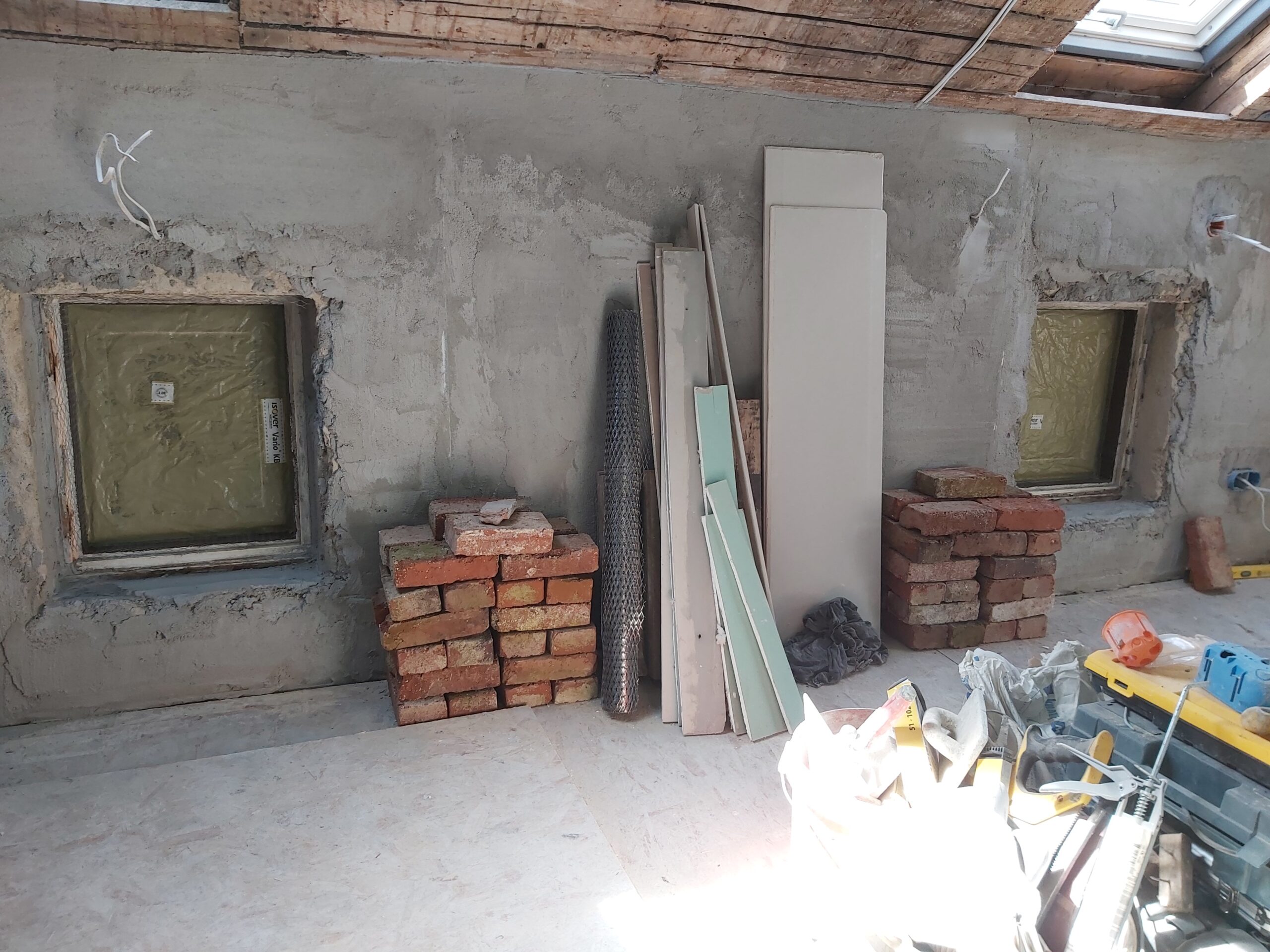 Ściana poddasza w trakcie remontu z dwiema wnękami okiennymi, ułożonymi cegłami, płytami gipsowymi i materiałami budowlanymi przygotowanymi do dalszych prac wykończeniowych.