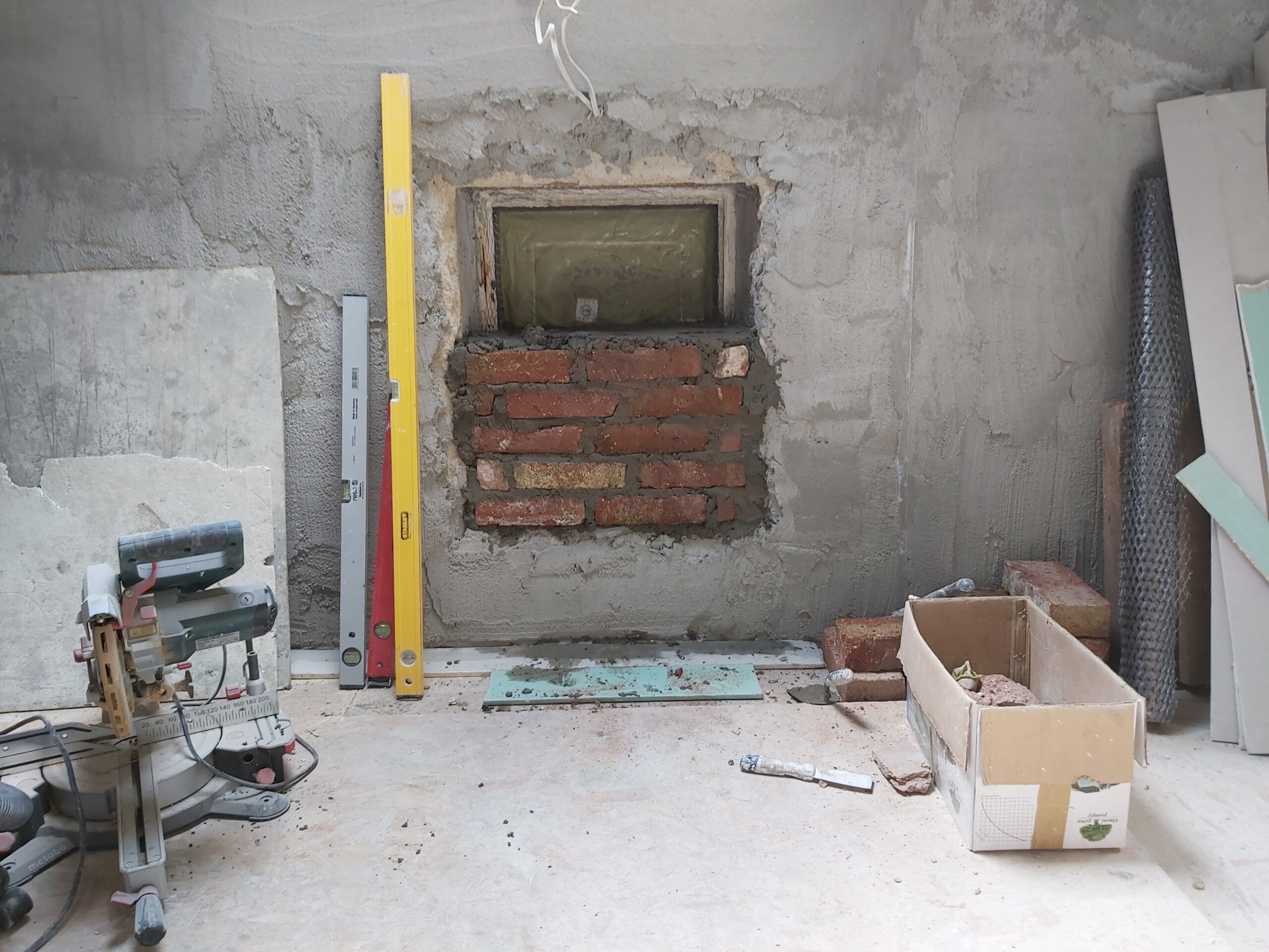 Zamurowywana cegłą wnęka w ścianie podczas remontu wnętrza, widoczna zaprawa murarska, poziomice, karton z materiałami oraz narzędzia budowlane na podłodze.