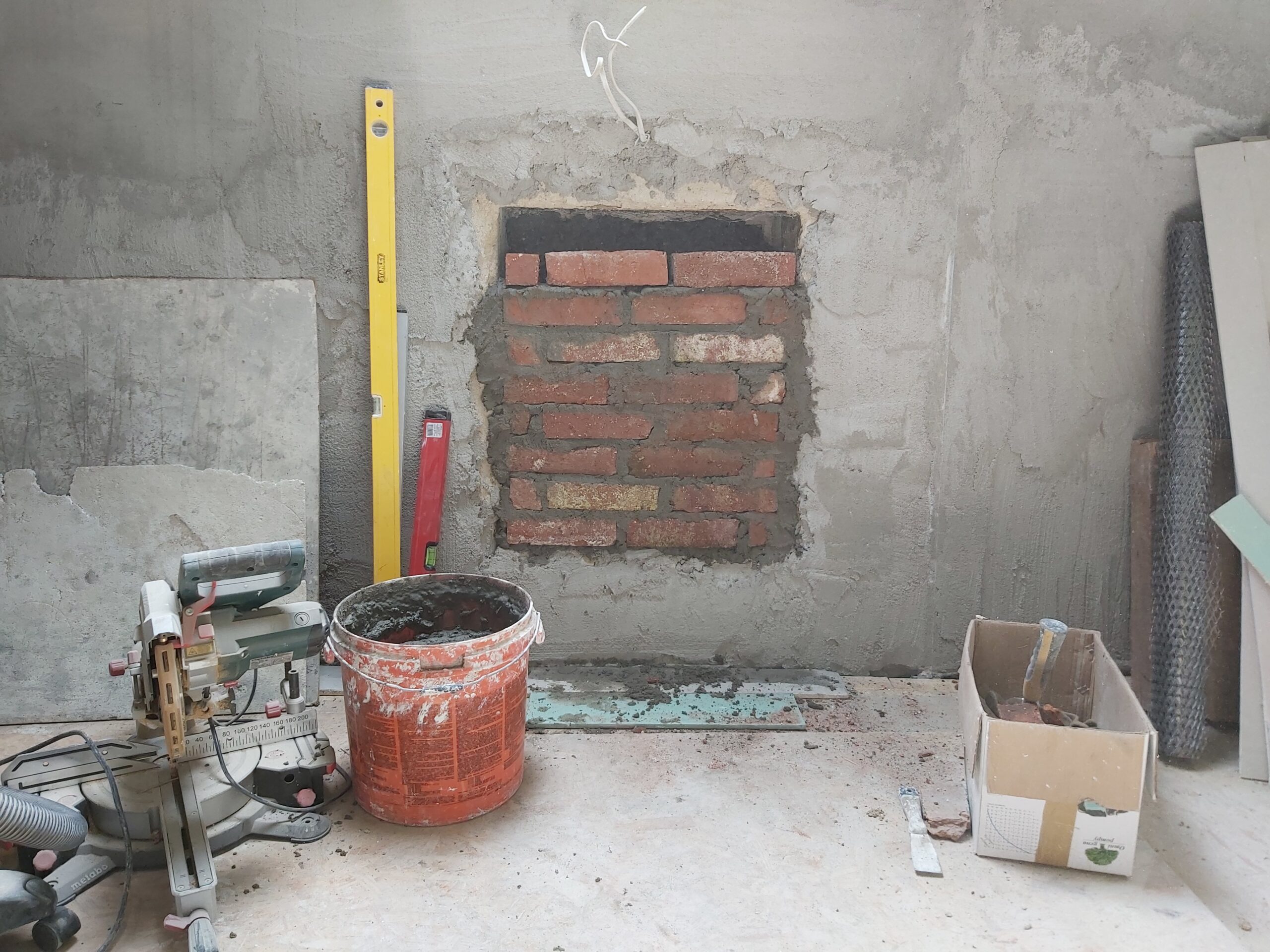 Wnętrze pomieszczenia podczas remontu – zamurowana cegłą wnęka w ścianie, świeża zaprawa murarska, wiadro z zaprawą, poziomica, karton z materiałami oraz narzędzia budowlane.
