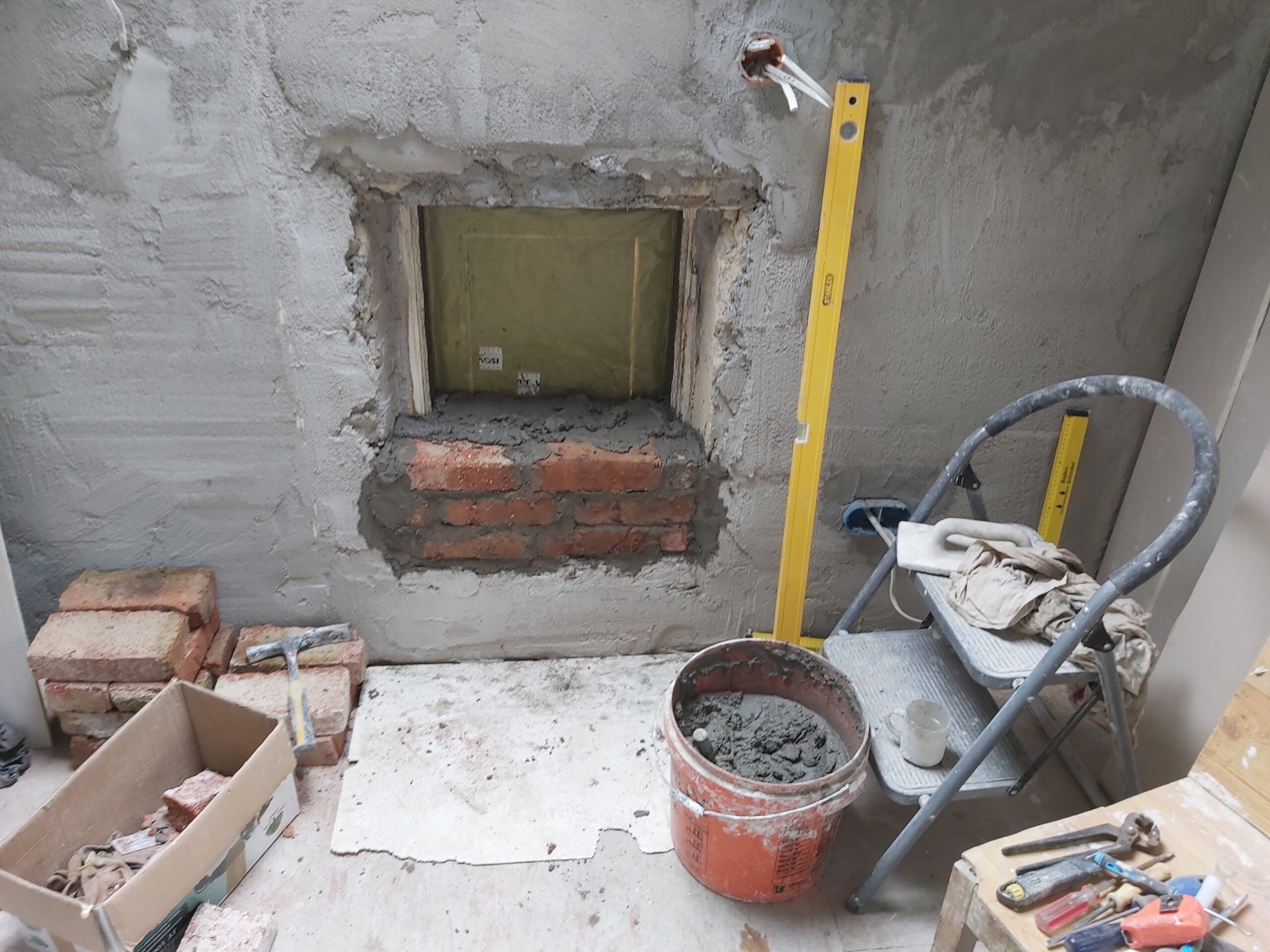 Ściana wewnętrzna podczas remontu z częściowo zamurowaną wnęką, widoczne cegły, świeża zaprawa murarska, wiadro z zaprawą, poziomica oraz narzędzia budowlane.