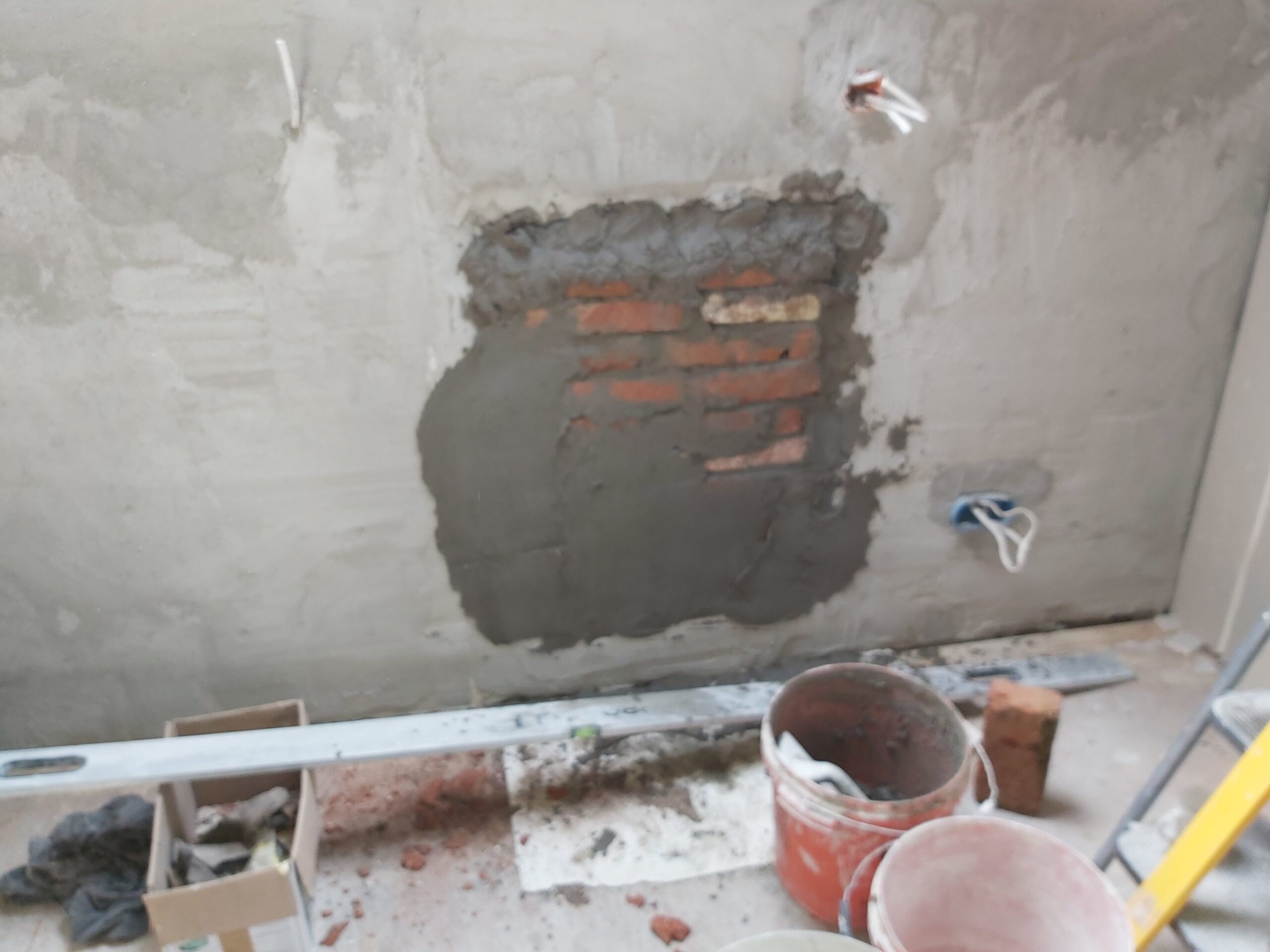 Ściana wewnętrzna w trakcie remontu z uzupełnianym ubytkiem, widoczne odsłonięte cegły, świeża zaprawa murarska, wiadra z zaprawą oraz elementy instalacji elektrycznej.