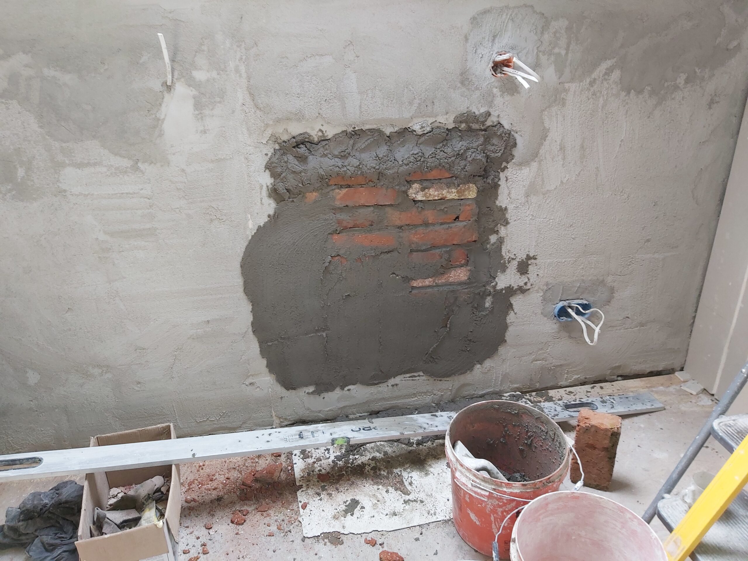 Fragment ściany wewnętrznej podczas remontu, z odsłoniętą cegłą i świeżą zaprawą murarską, widoczne wiadra z zaprawą, przewody elektryczne oraz puszka instalacyjna.