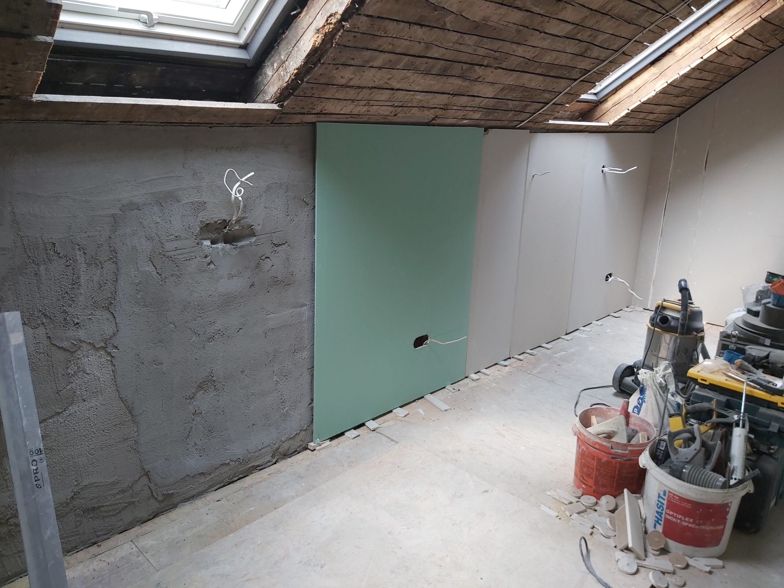 Poddasze w trakcie remontu, ściany częściowo pokryte płytami gipsowo-kartonowymi, widoczna izolacja, instalacja elektryczna oraz narzędzia budowlane na podłodze.