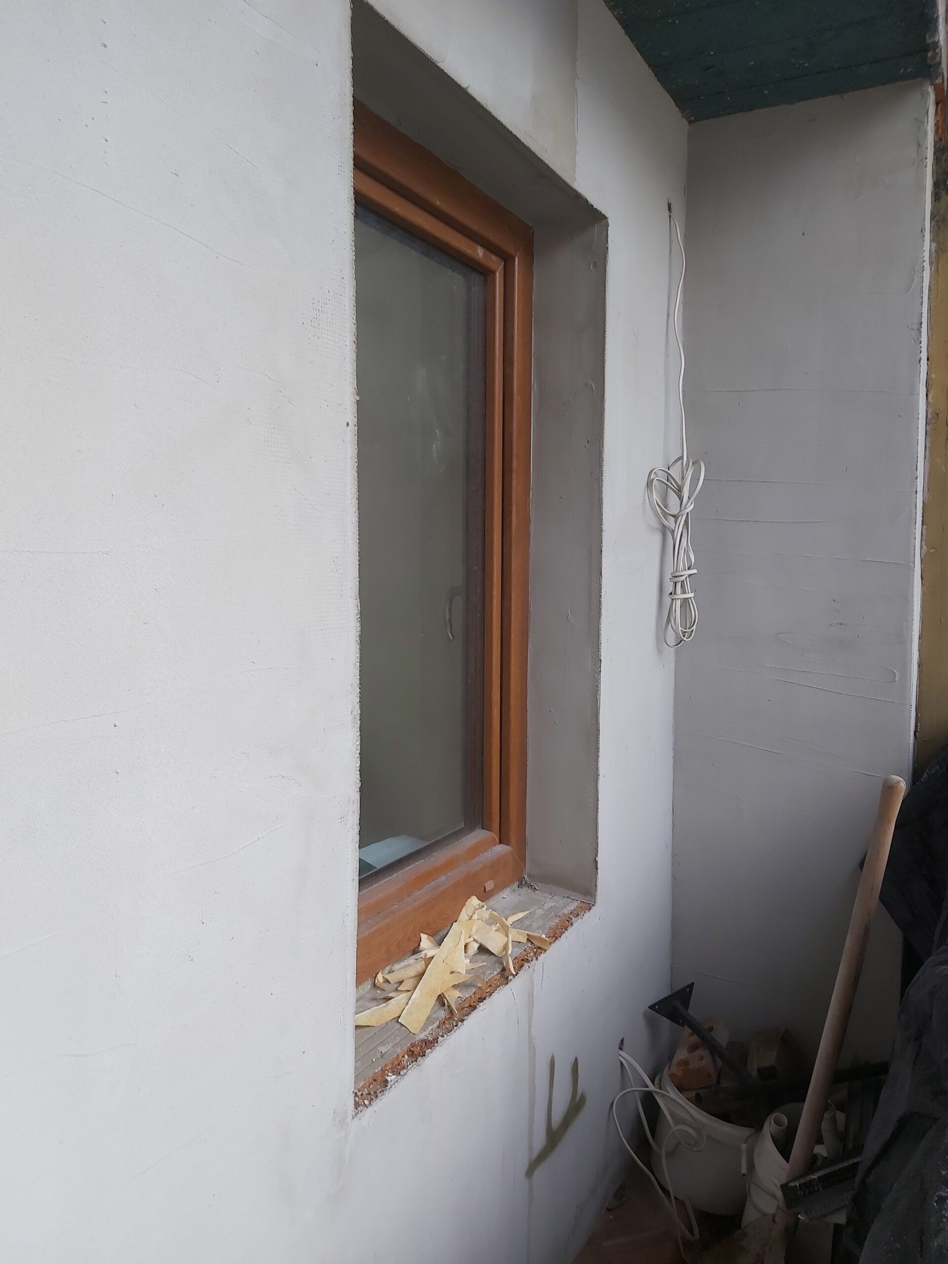 Ościeże okna z zamontowaną drewnianą ramą, świeżo wykończone powierzchnie ścian oraz widoczne przewody i materiały używane podczas prac wykończeniowych.