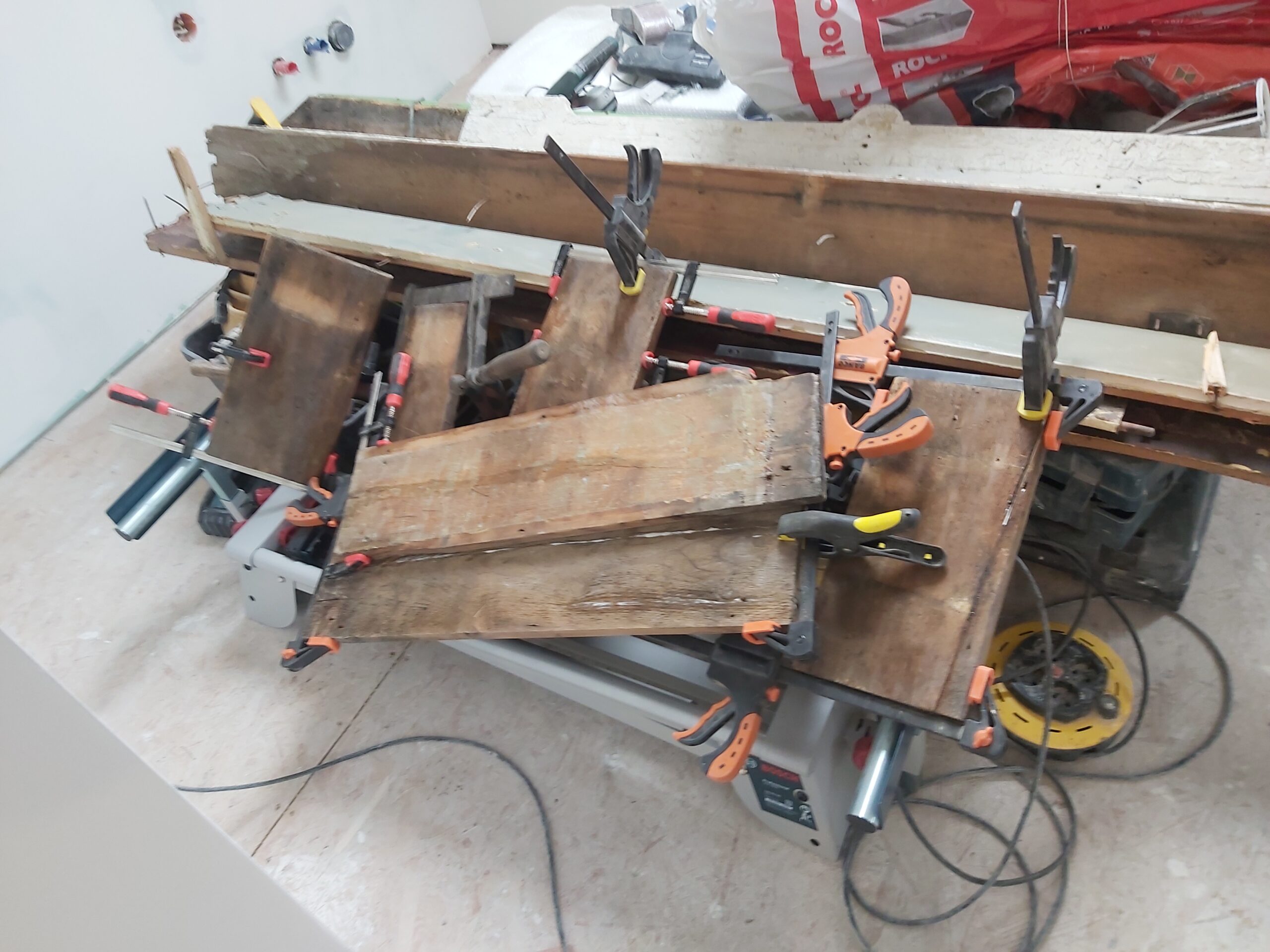 Elementy drewniane łączone i dociskane ściskami stolarskimi na stanowisku roboczym, widoczne narzędzia oraz przygotowanie materiału do dalszego montażu.