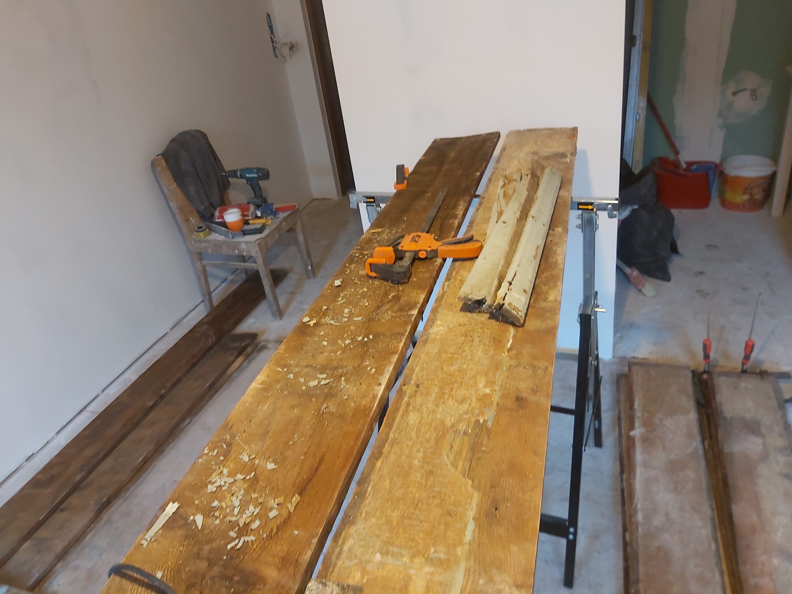 Stanowisko robocze z drewnianymi deskami w trakcie obróbki, widoczne ściski stolarskie, narzędzia oraz przygotowanie materiału do dalszego montażu.