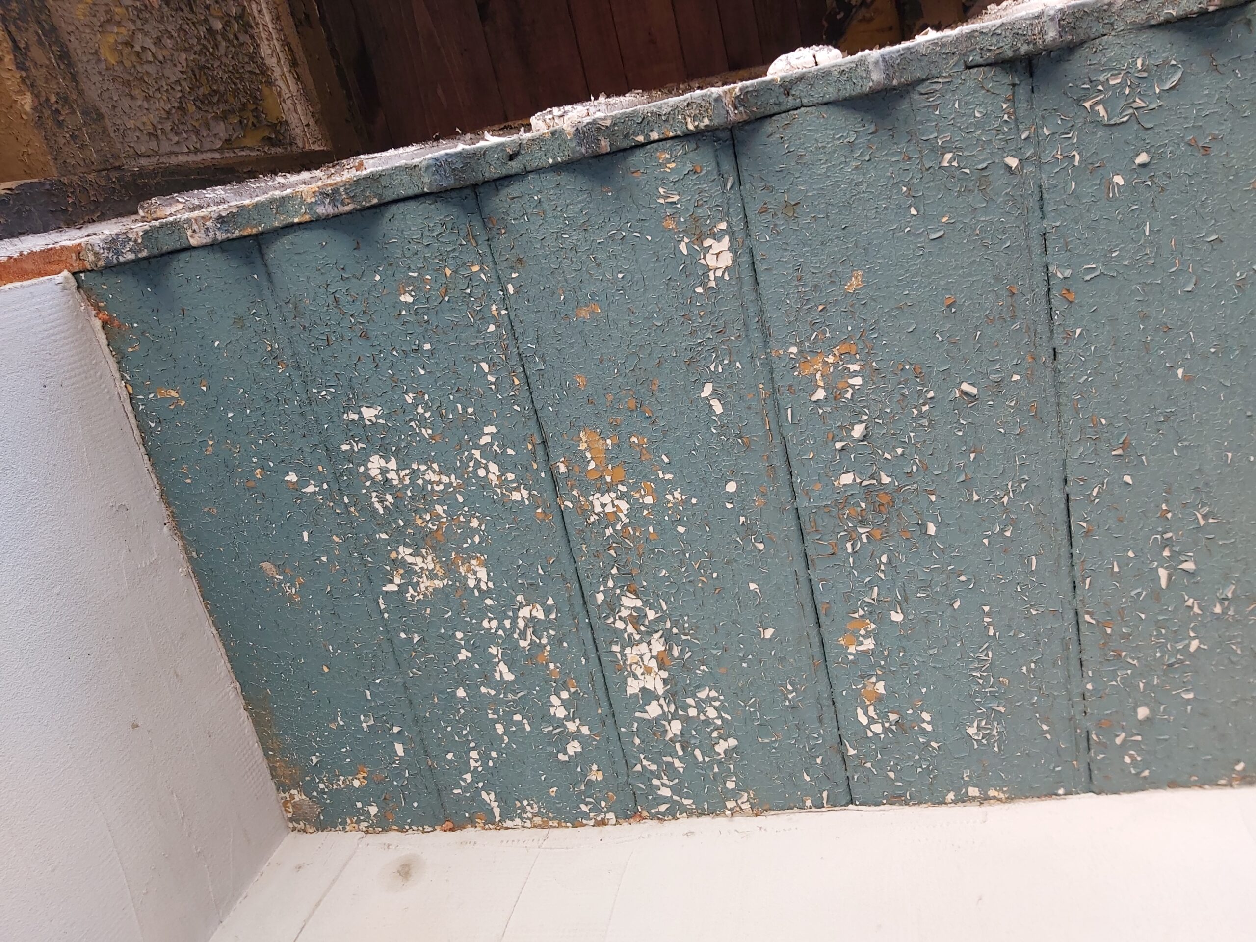 Fragment sufitu z widoczną starą, łuszczącą się powłoką malarską na drewnianych deskach, przygotowywany do dalszych prac remontowych.