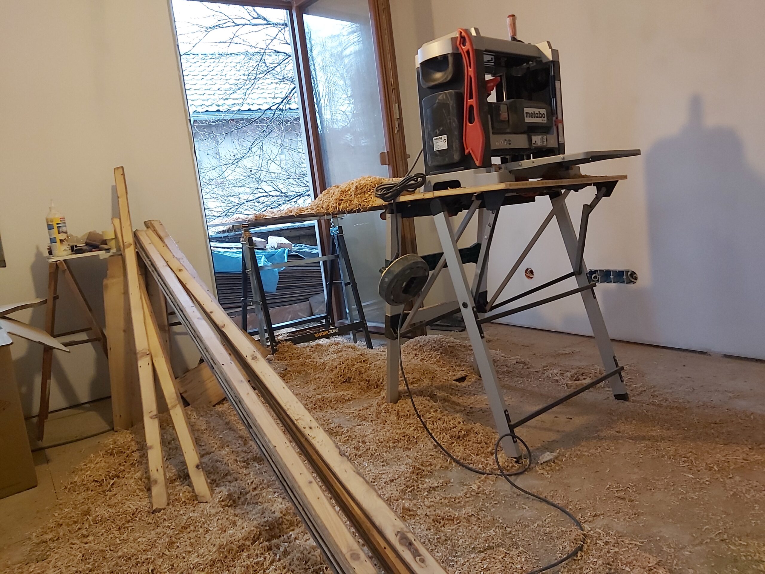Pomieszczenie w trakcie remontu z pilarką stołową, obrabianymi elementami drewnianymi oraz trocinami na podłodze, widoczne przygotowanie materiałów do montażu.