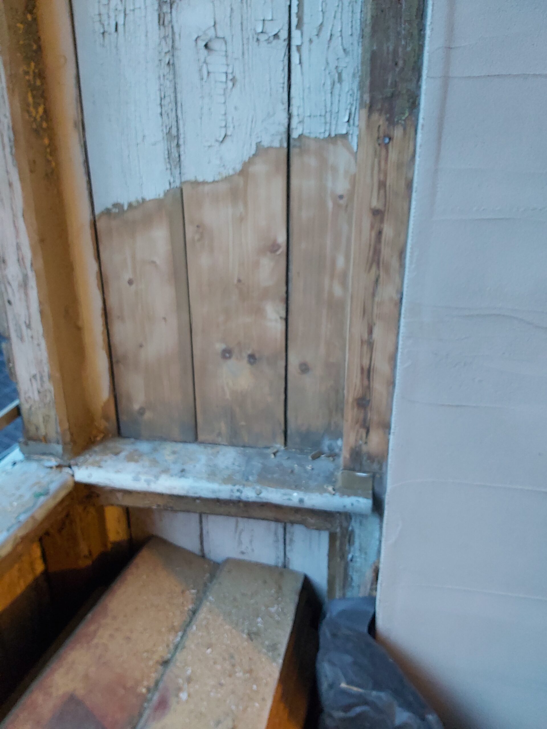 Zbliżenie na ościeże okna z odsłoniętymi drewnianymi deskami i fragmentami starej farby, widoczne przygotowanie do dalszych prac wykończeniowych.