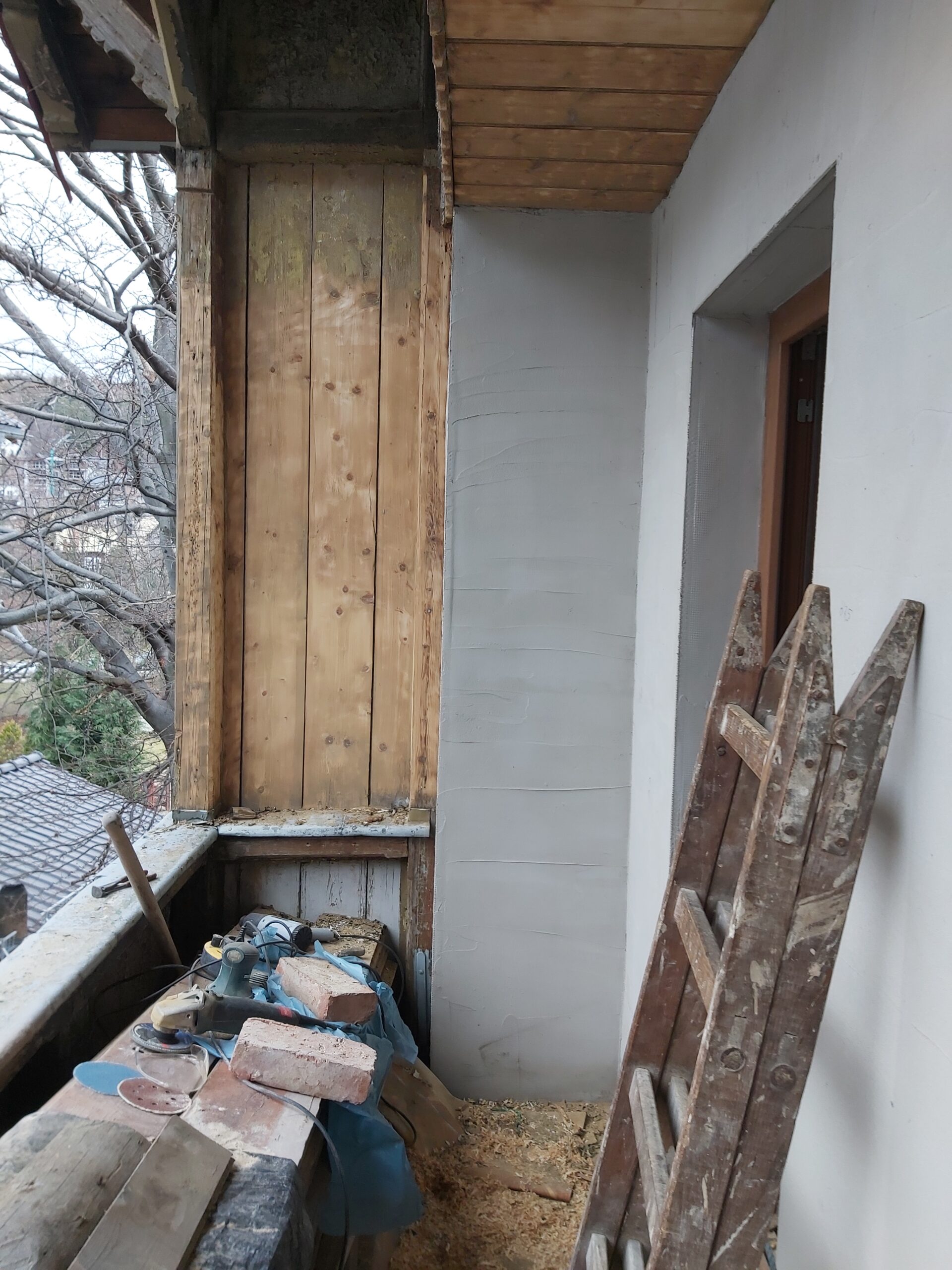 Balkon podczas remontu, widoczny drewniany słup konstrukcyjny, fragment sufitu z desek, narzędzia i materiały budowlane przygotowane do dalszych prac.