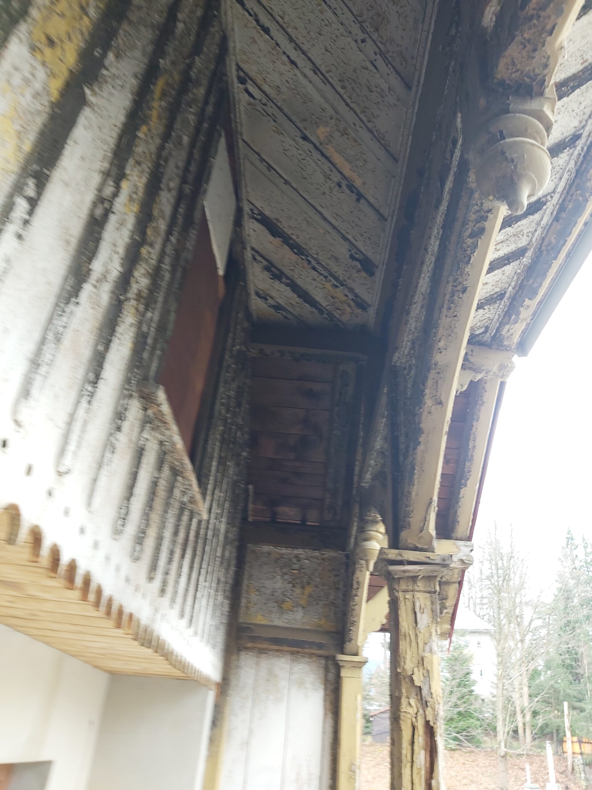 Widok od spodu na drewniany balkon z ozdobnymi wspornikami, łuszczącą się farbą i widocznymi uszkodzeniami drewna przed pracami renowacyjnymi.