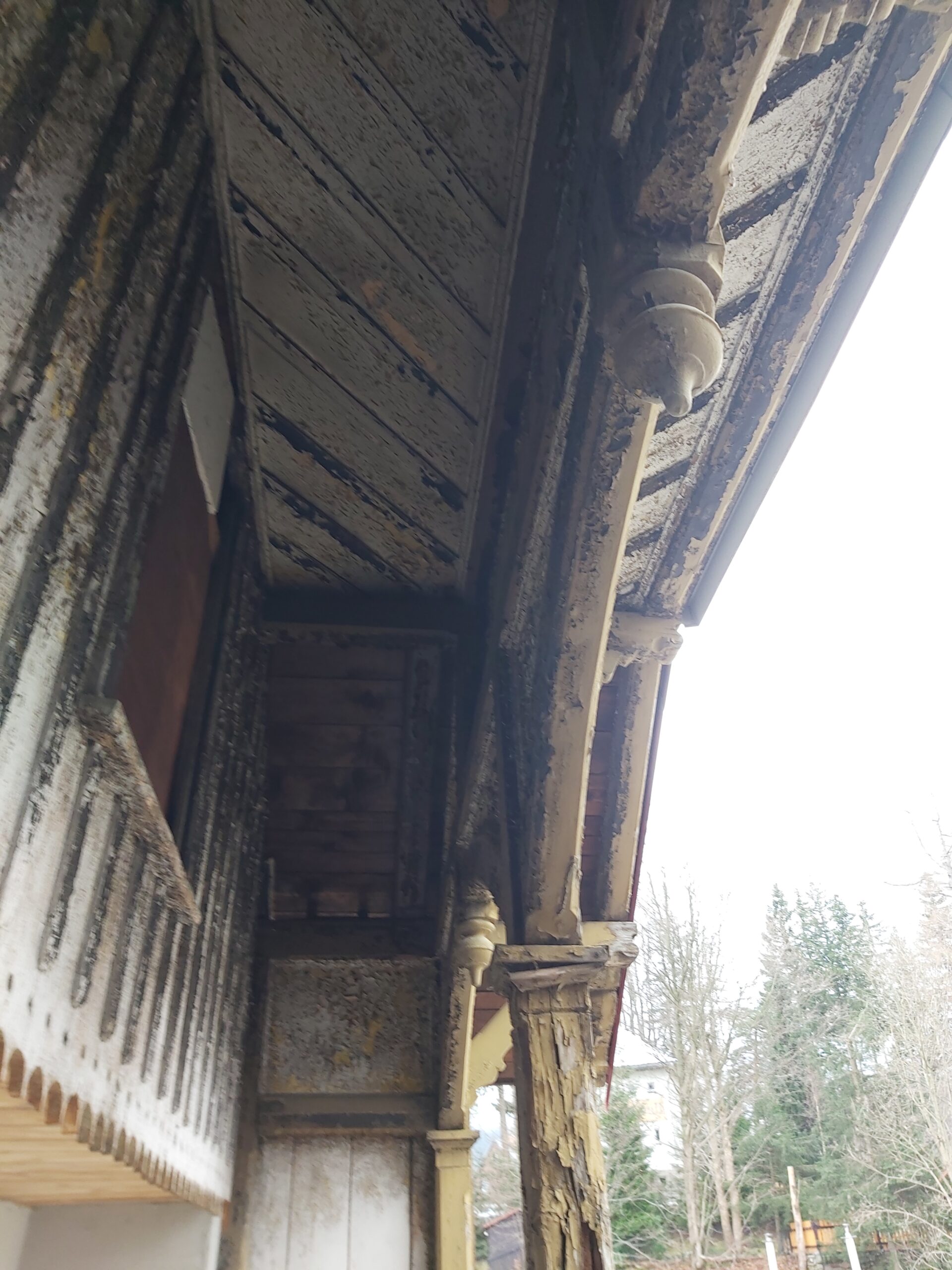 Stara drewniana konstrukcja balkonu z dekoracyjnymi wspornikami i łuszczącą się farbą, ukazująca zaawansowane zużycie drewna przed renowacją.