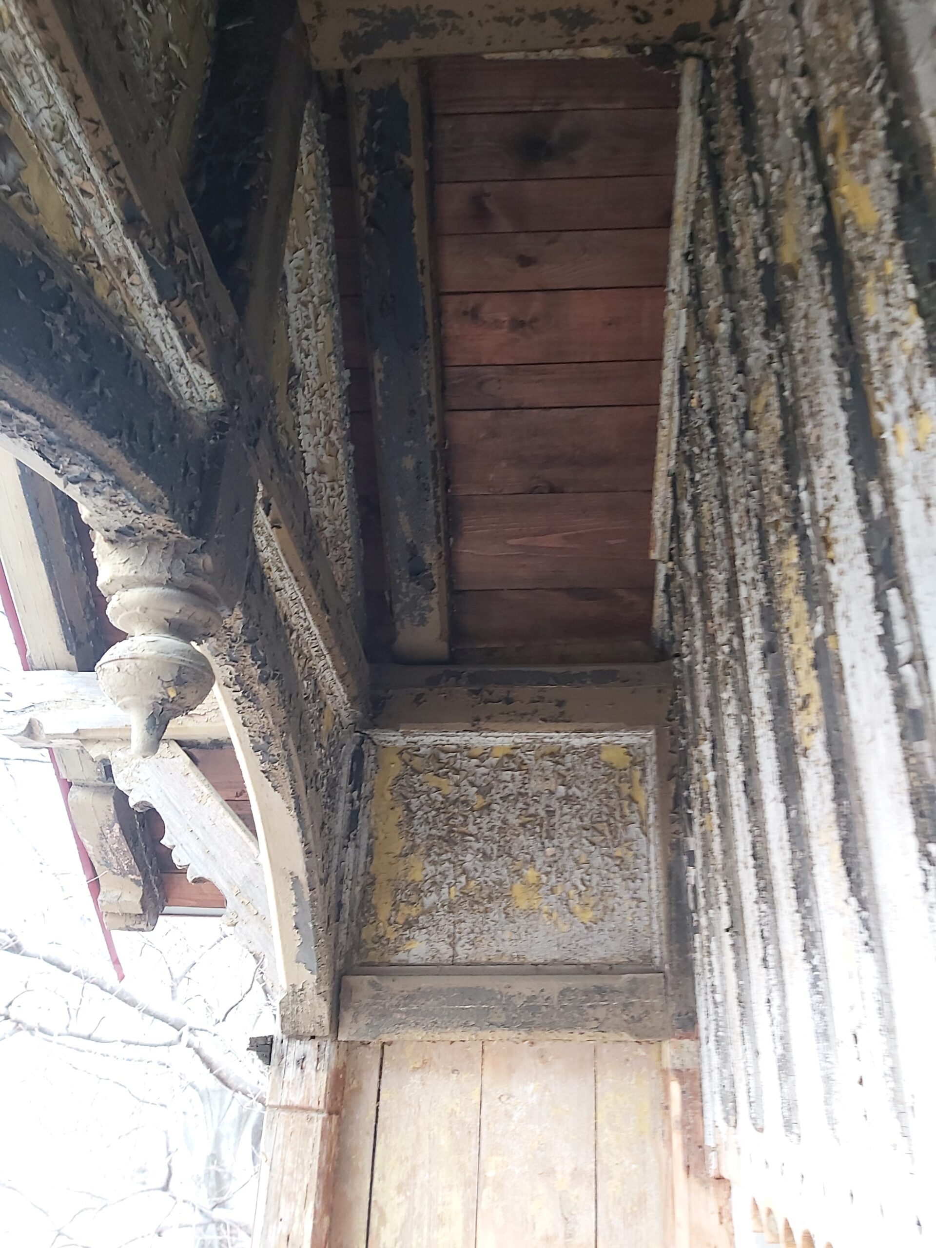 Zbliżenie na starą drewnianą konstrukcję balkonu z łuszczącą się farbą, widocznymi belkami i dekoracyjnym wspornikiem podczas remontu.