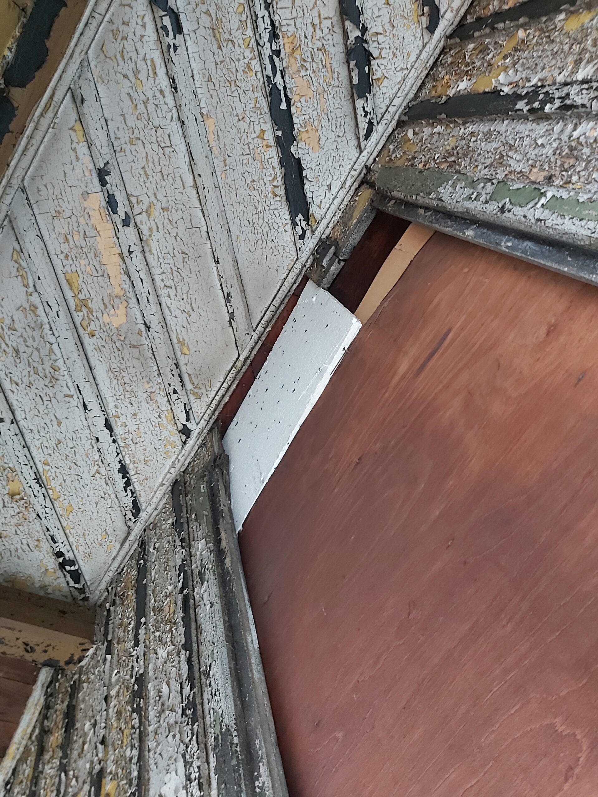 Zbliżenie na starą drewnianą okładzinę balkonu z łuszczącą się farbą oraz fragmentem nowej płyty zabezpieczającej konstrukcję w trakcie remontu.