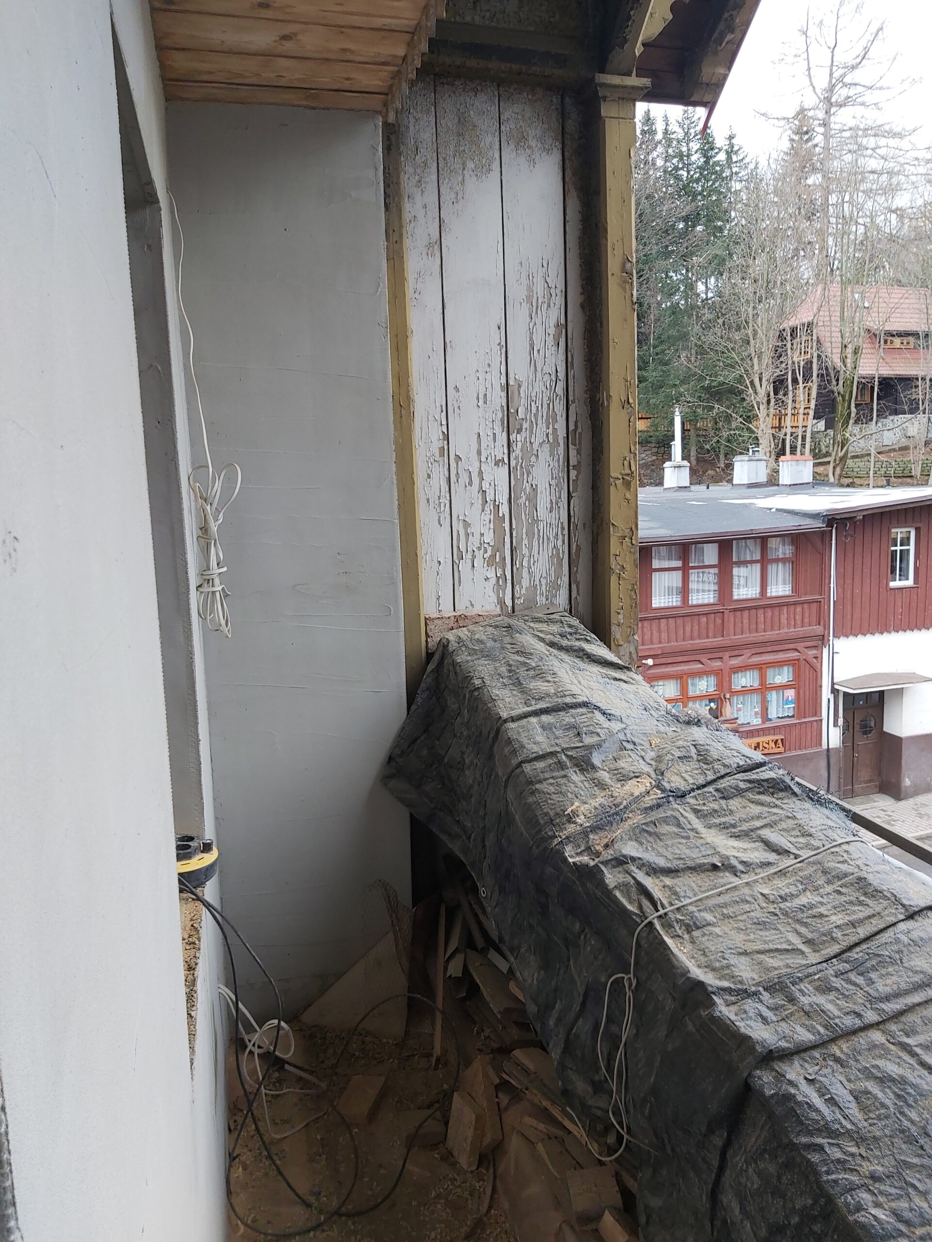 Ściana balkonu z odkrytą, starą drewnianą okładziną, fragmentem konstrukcji osłoniętym plandeką oraz przewodami i narzędziami w trakcie remontu.