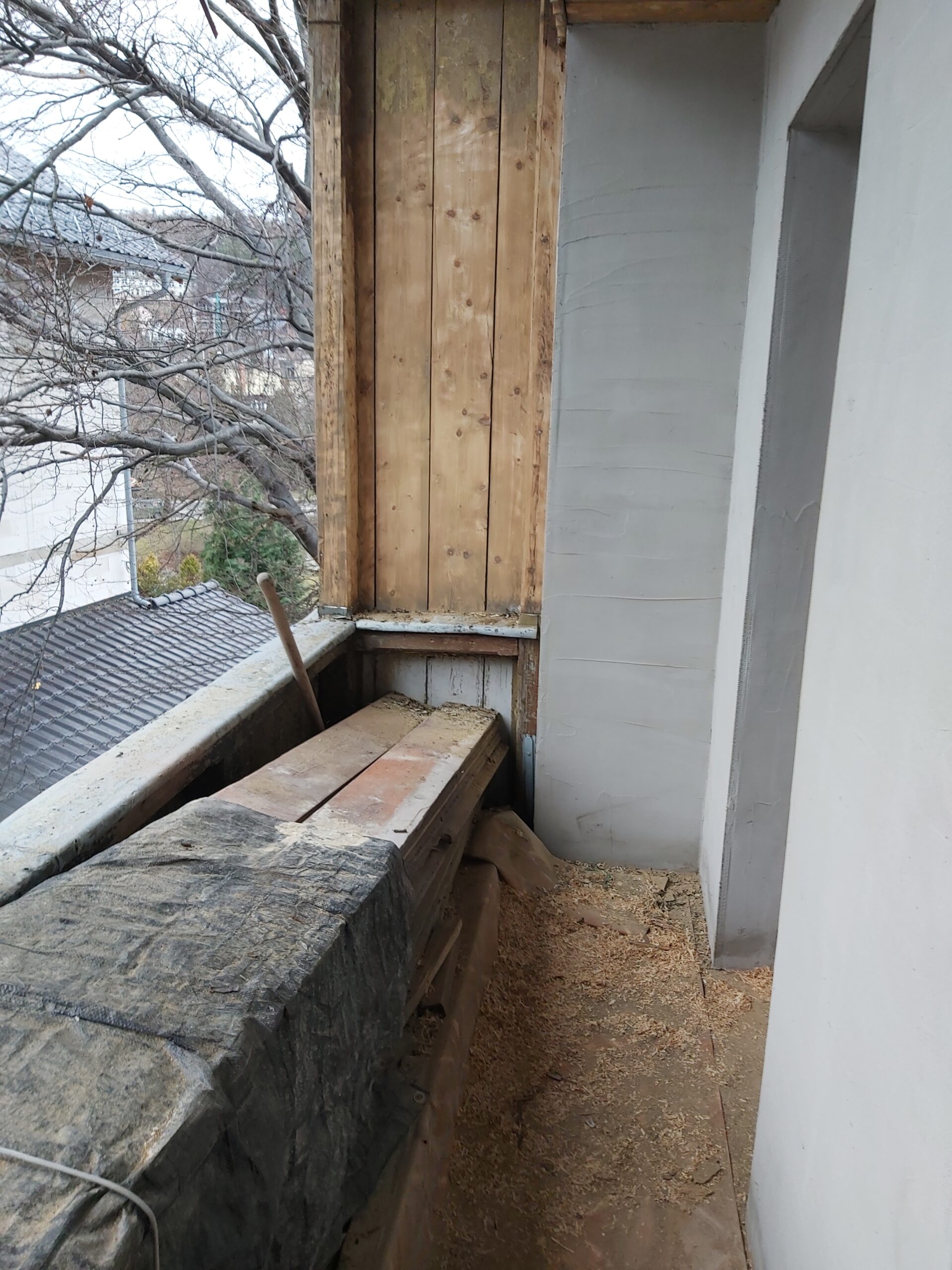 Narożnik balkonu z odsłoniętą drewnianą konstrukcją, widocznymi deskami, prowizorycznym zabezpieczeniem i przygotowaniem do dalszych prac remontowych.