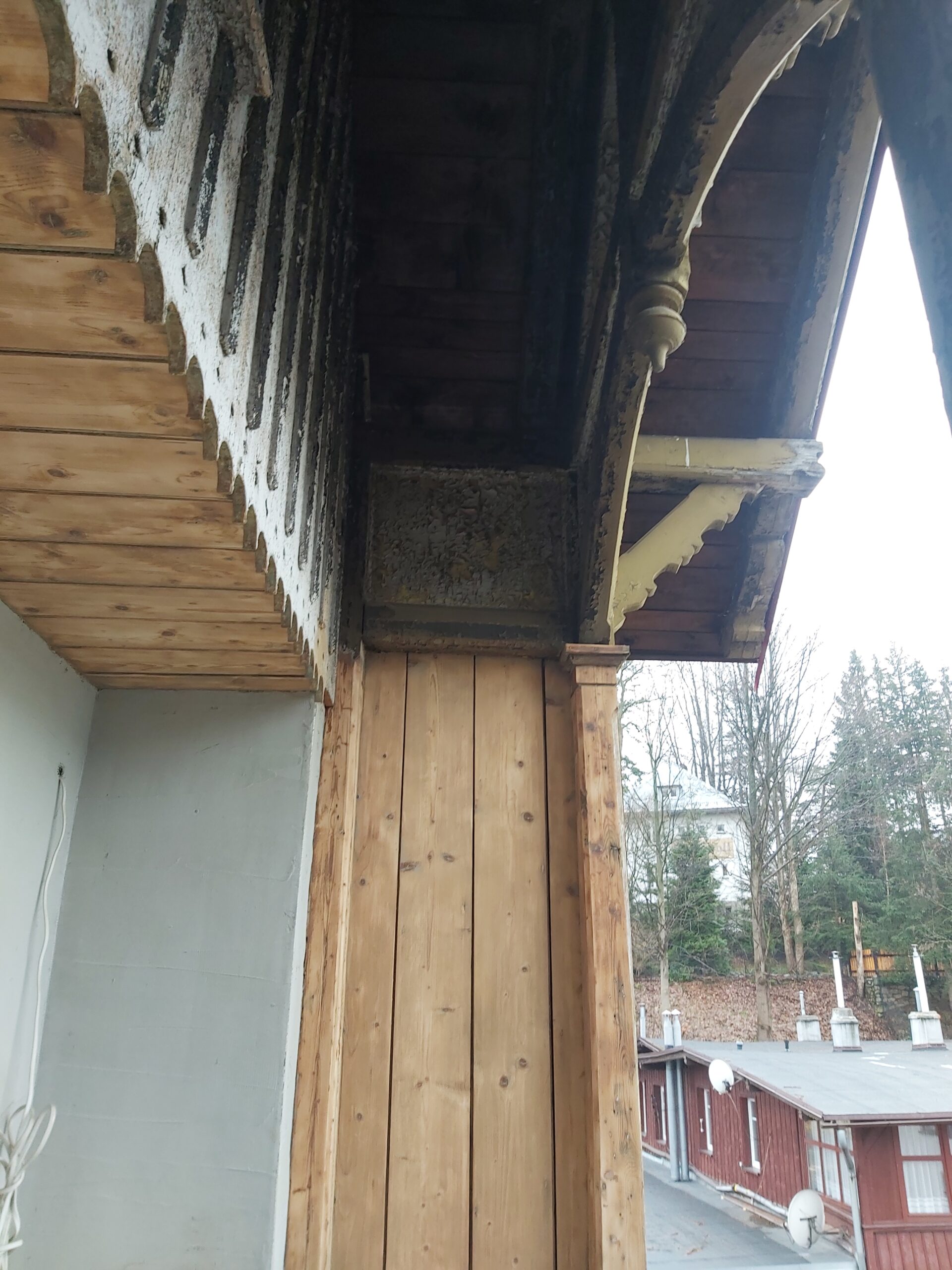 Widok od dołu na drewniany słup balkonu oraz okap z dekoracyjnymi wycięciami po oczyszczeniu starej farby, podczas prac remontowych.