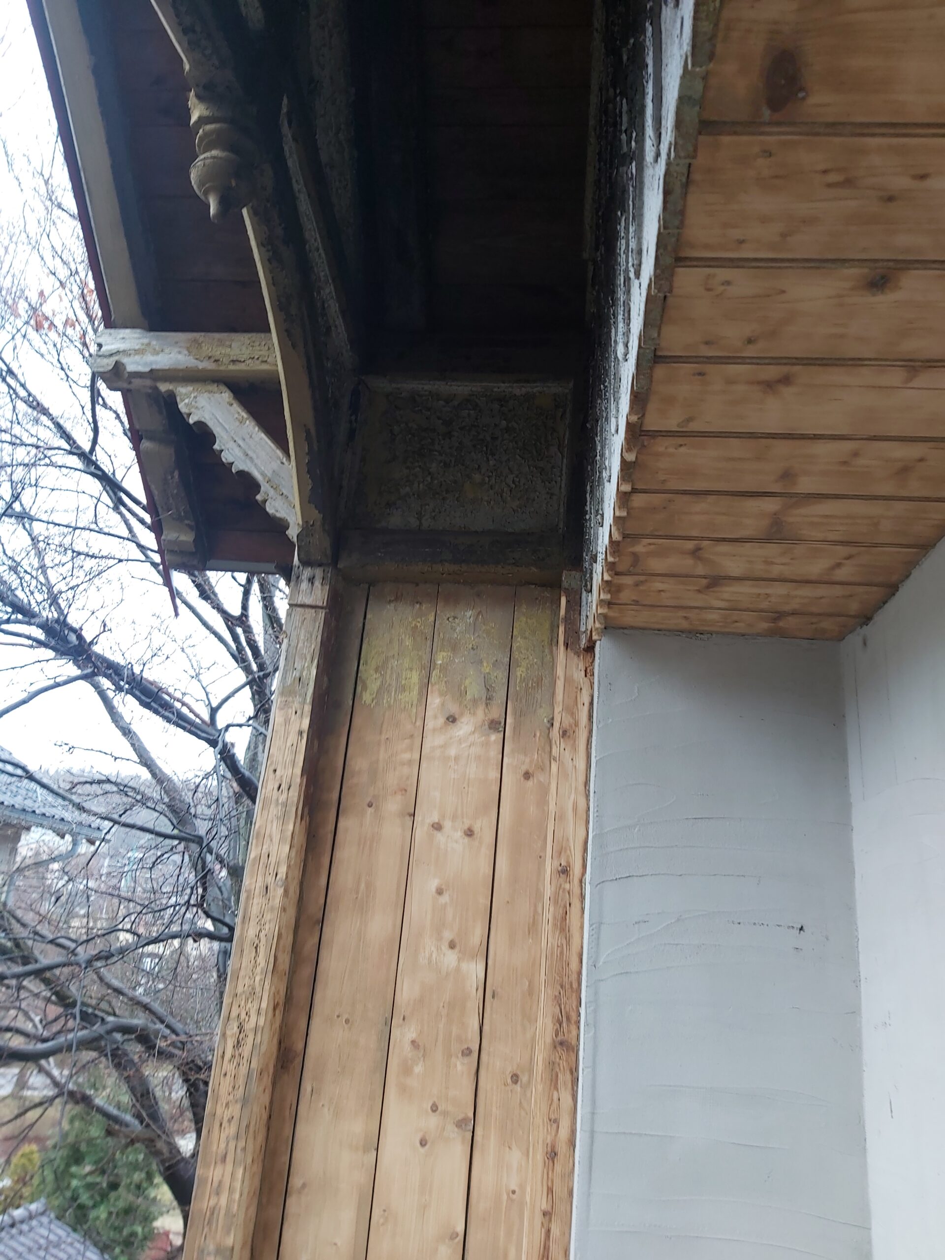 Oczyszczony fragment drewnianej konstrukcji balkonu z widocznym słupem, belką stropową i spodem okapu w trakcie remontu.