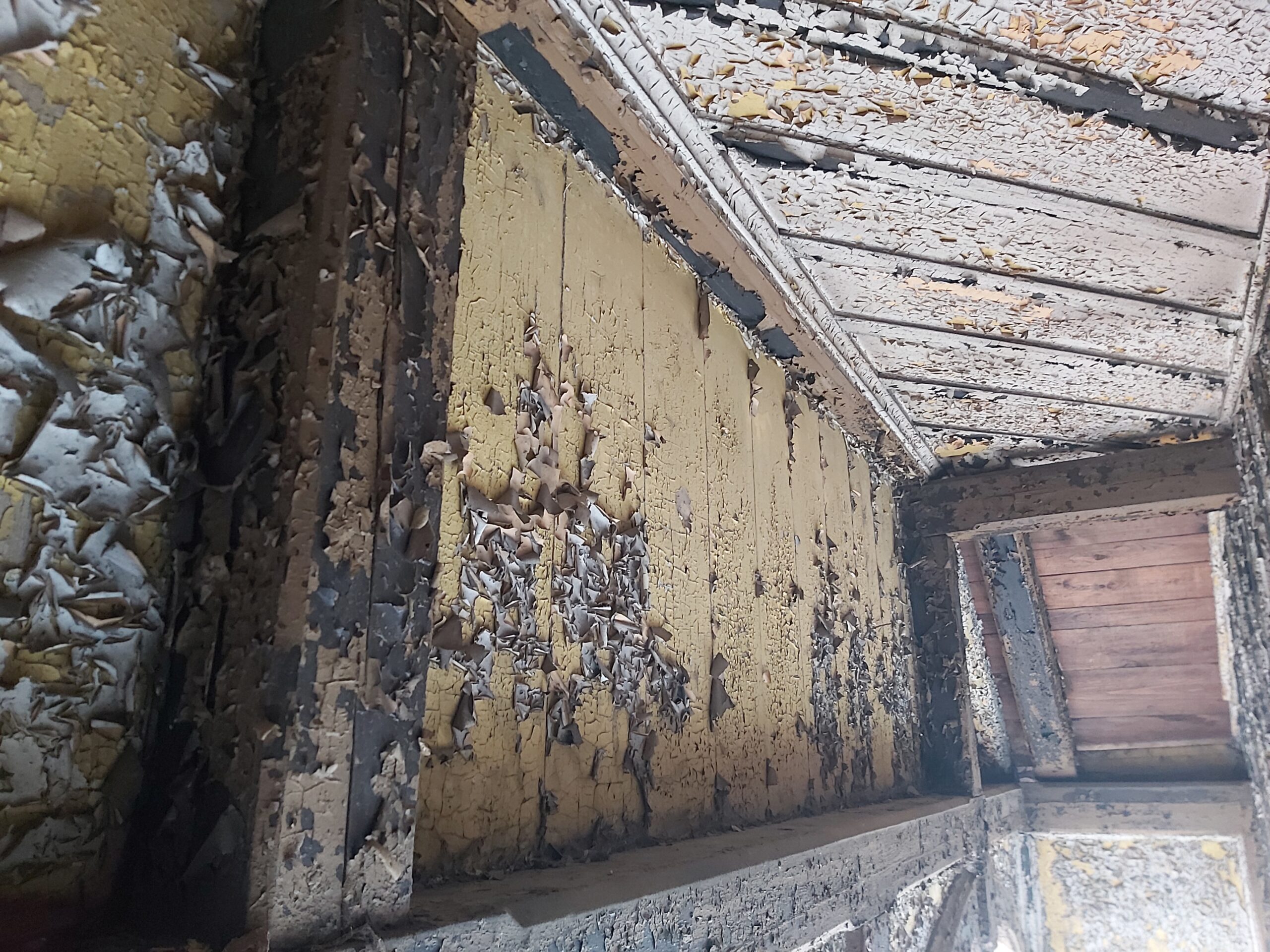 Fragment drewnianej ściany balkonu z mocno łuszczącą się starą farbą i widocznymi śladami wieloletniej degradacji podczas remontu.