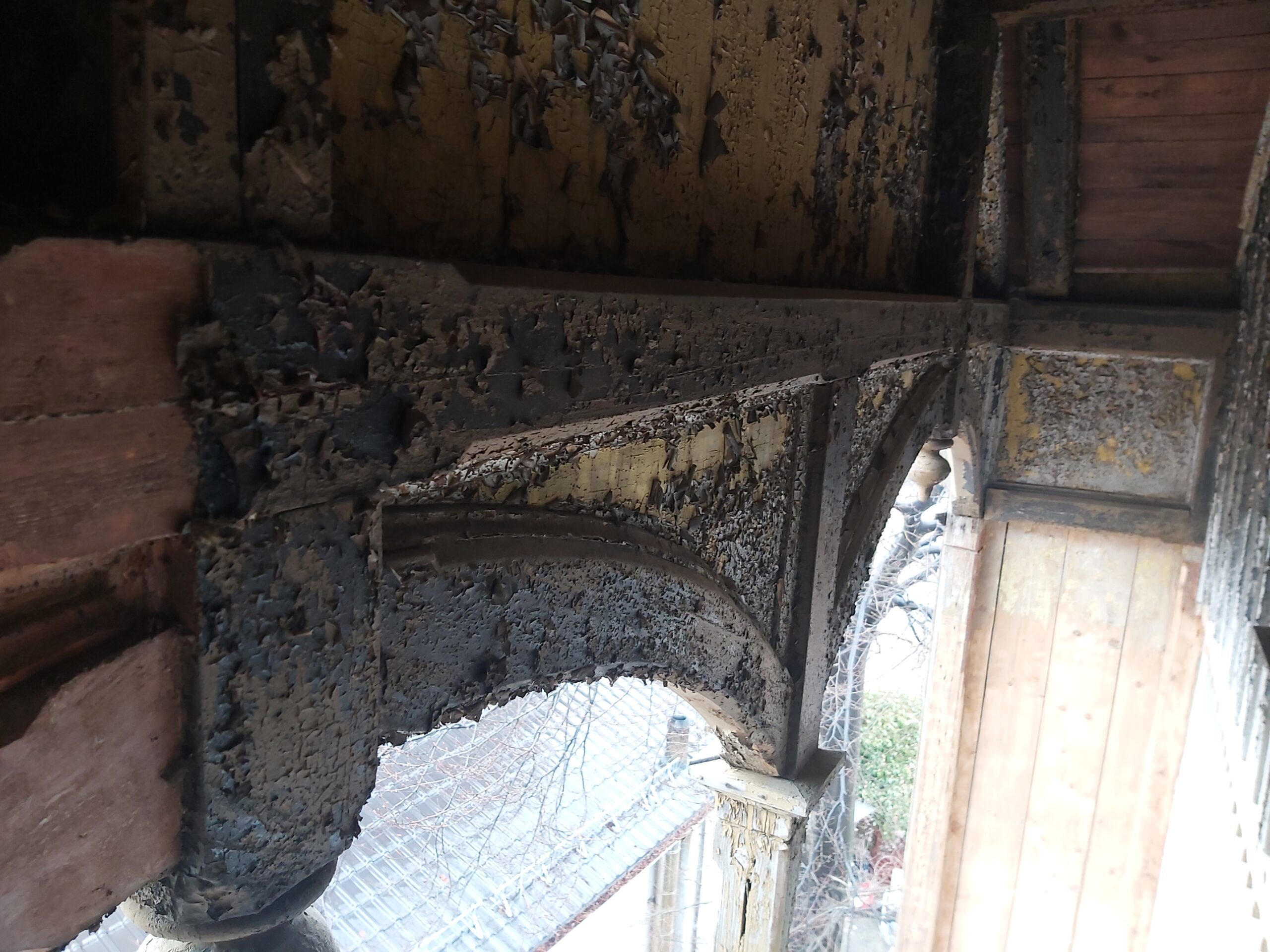 Drewniany łuk balkonu z widoczną, łuszczącą się farbą i oznakami wieloletniego zużycia, fragment odkryty w trakcie remontu konstrukcji.