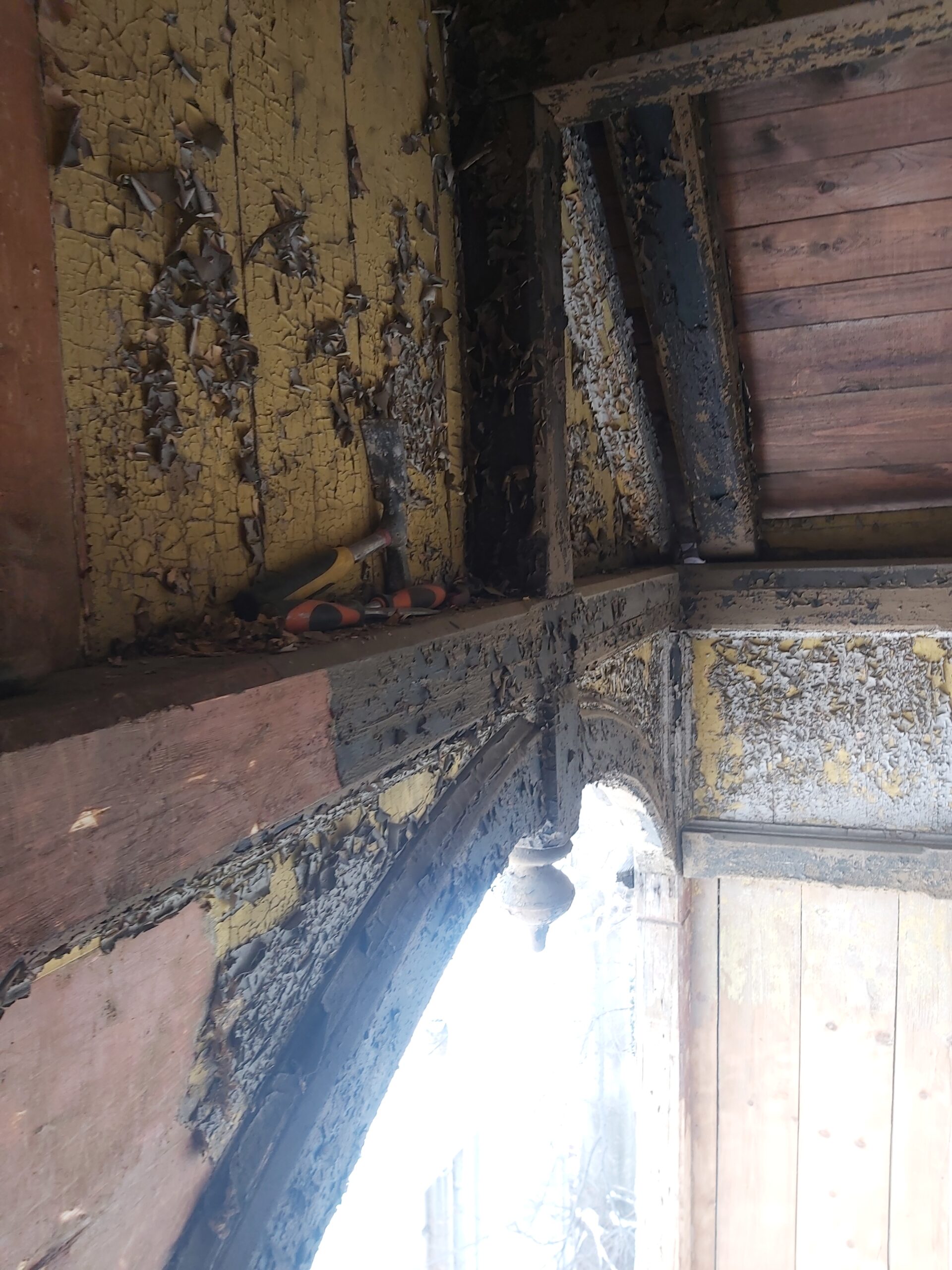 Narożnik balkonu z odsłoniętym drewnem i starą, łuszczącą się farbą, widoczne ślady wilgoci i degradacji konstrukcji podczas remontu.