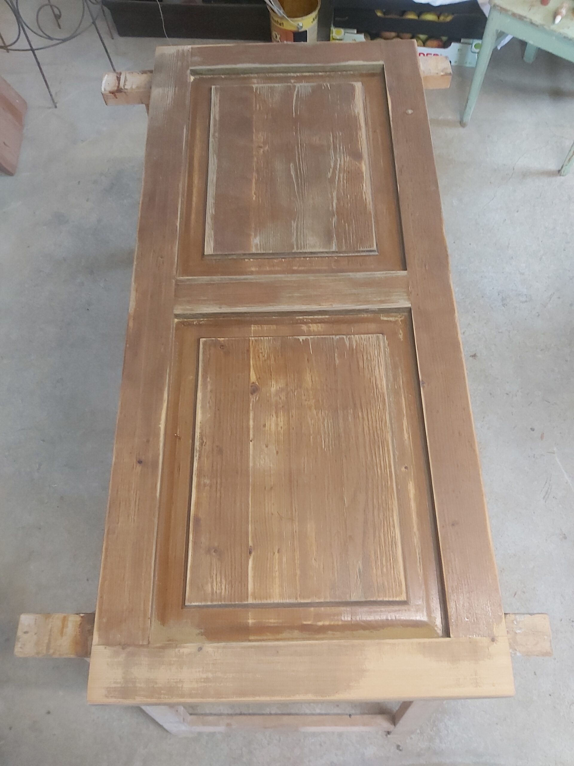 Pojedynczy drewniany front meblowy ułożony poziomo na kozłach, z widocznymi śladami zużycia, przetarciami i nierówną powierzchnią po wcześniejszym wykończeniu.