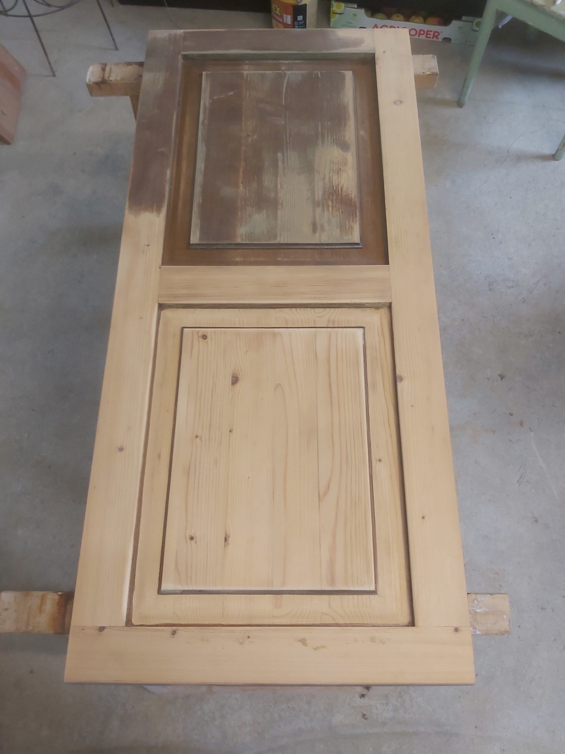 Drewniane drzwi ułożone poziomo na kozłach, z widocznymi śladami szlifowania i częściowo usuniętej starej powłoki w górnym panelu.