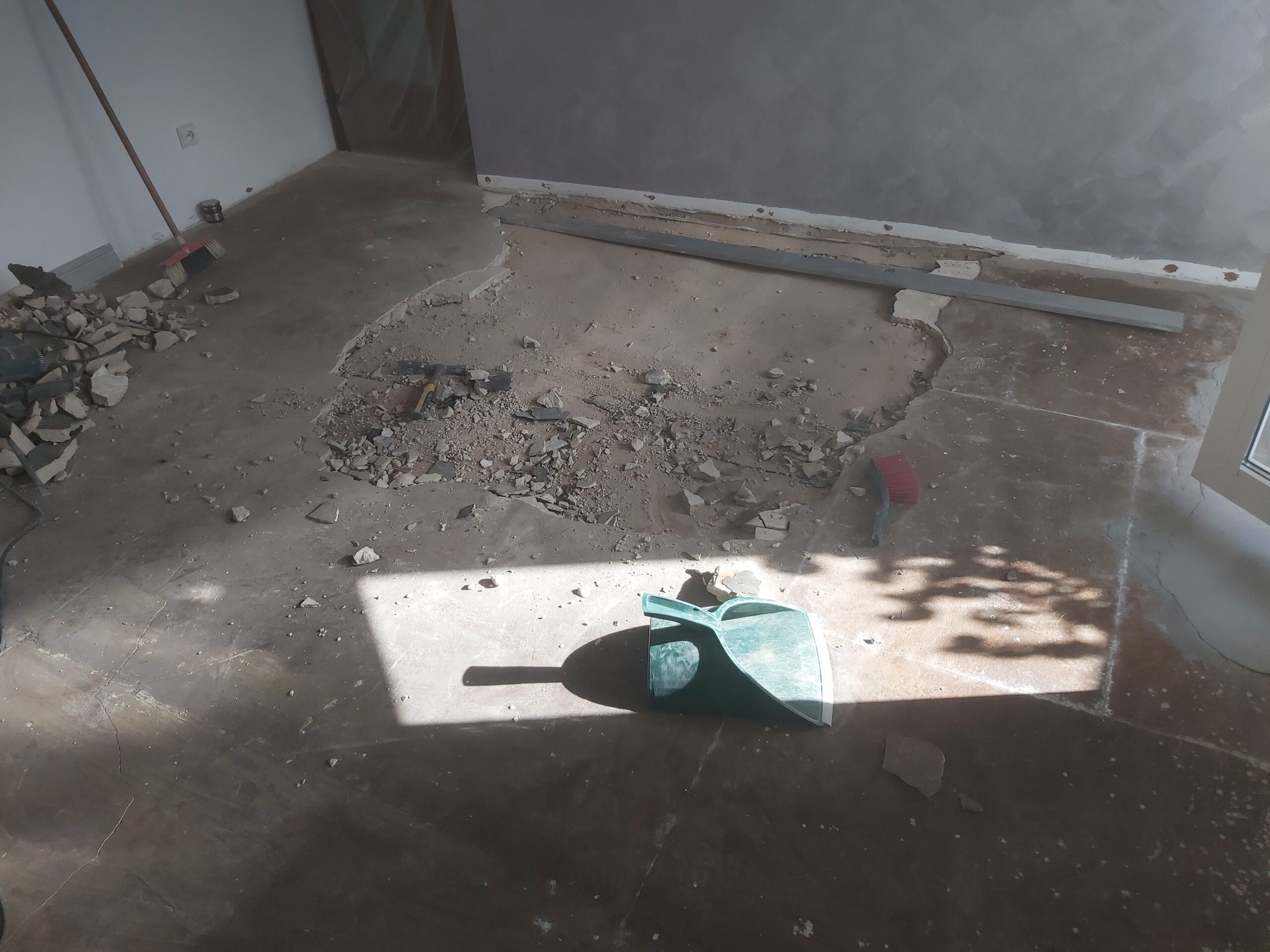 Podłoga podczas prac remontowych z wykutym fragmentem starej posadzki, rozsypanym gruzem oraz widoczną szuflą i narzędziami na tle pomieszczenia.