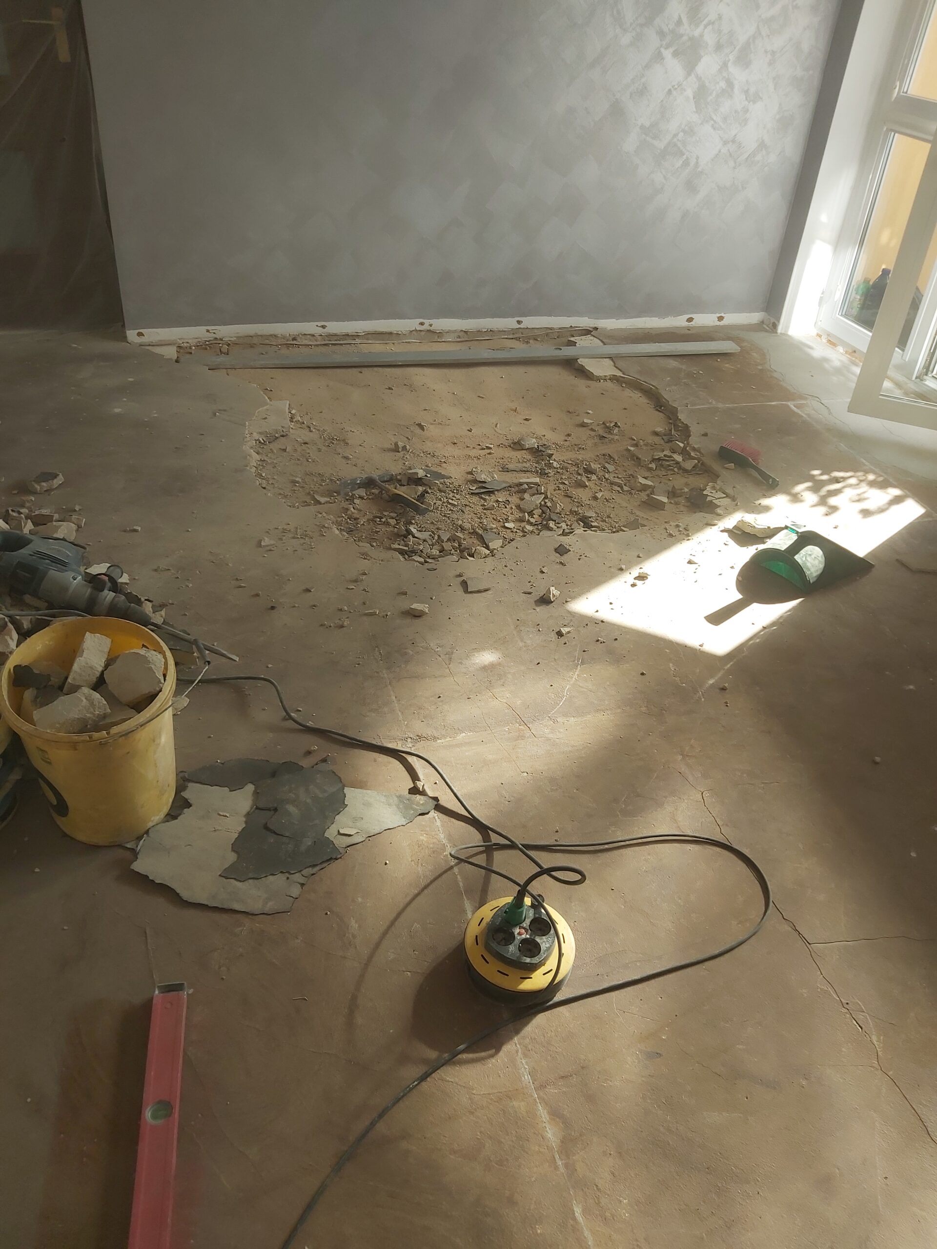 Podłoga w trakcie remontu z wykutym fragmentem starej posadzki, widoczne gruz, narzędzia oraz przedłużacz, przygotowanie powierzchni pod dalsze prace.