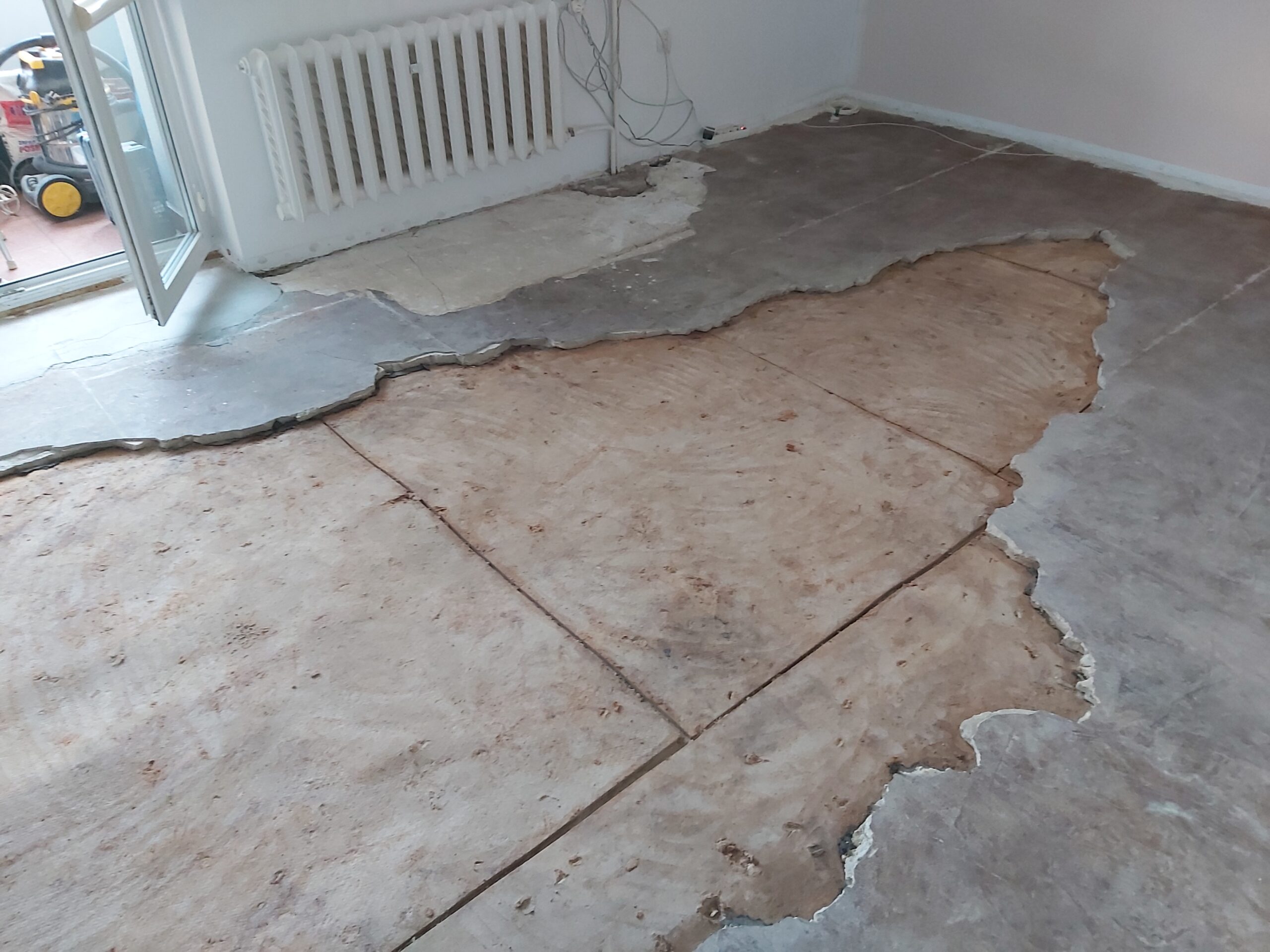 Fragment podłogi po usunięciu starej posadzki, z widoczną odsłoniętą płytą i nieregularnymi krawędziami pozostałych warstw w trakcie remontu.