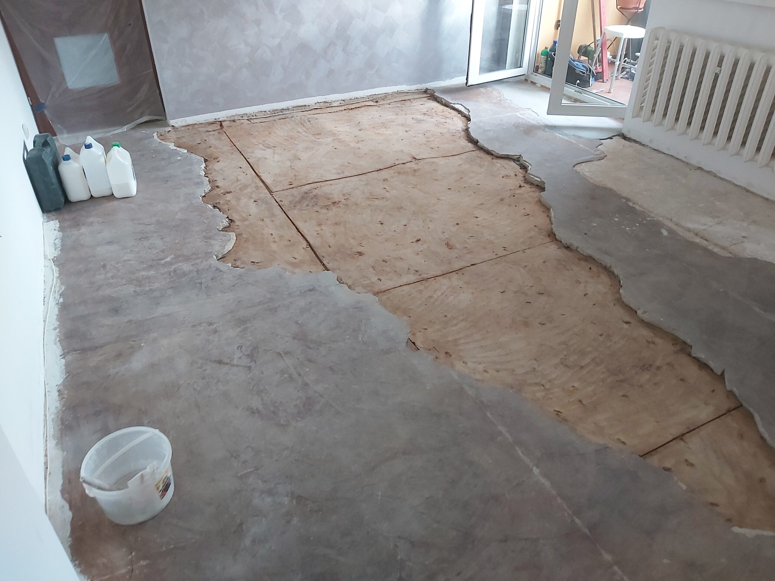 Pomieszczenie w trakcie remontu z usuniętą warstwą starej podłogi, odsłoniętym podłożem oraz widocznymi narzędziami i materiałami przygotowanymi do dalszych prac.