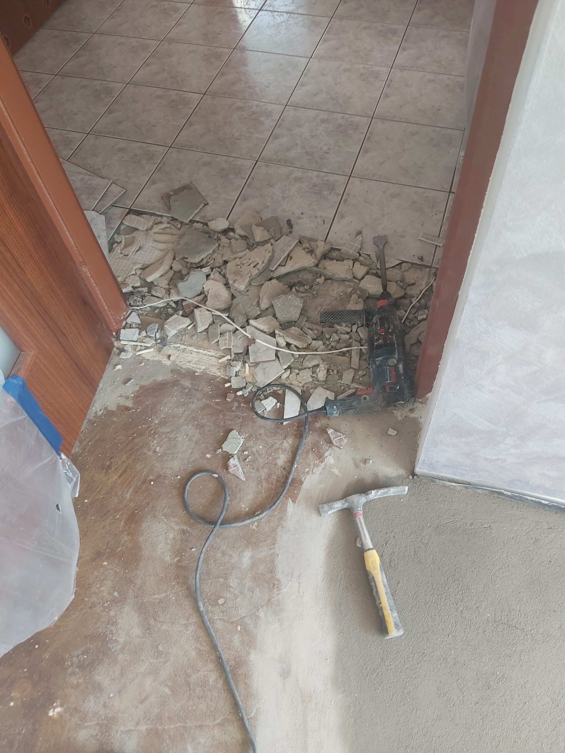 Próg drzwiowy w trakcie remontu, z rozkutymi płytkami ceramicznymi, gruzem, młotkiem oraz wiertarką leżącymi na podłodze.