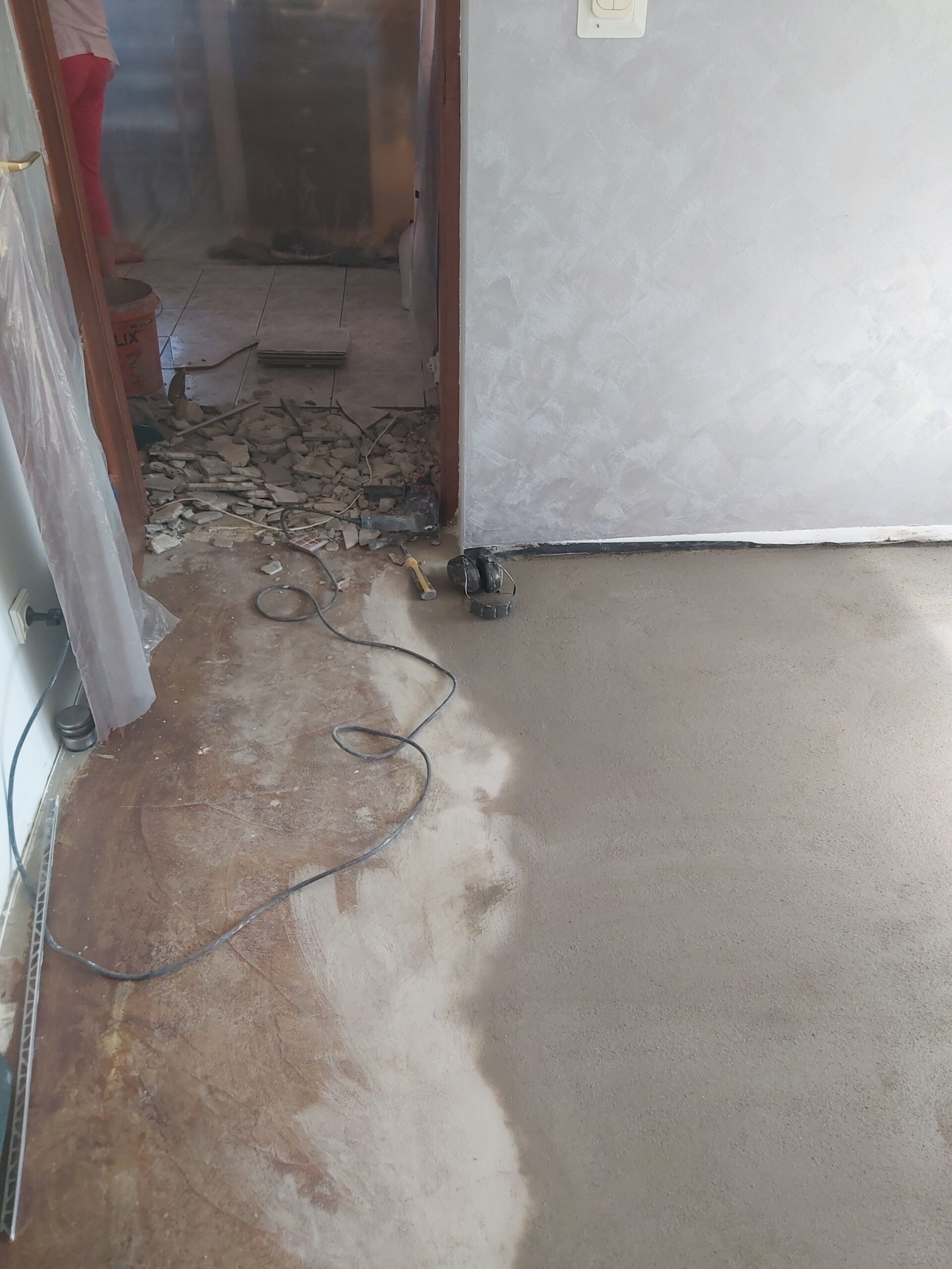Przejście między pomieszczeniami podczas remontu, z widocznym gruzem po skuciu starej posadzki, elektronarzędziami oraz świeżo wyrównaną wylewką.